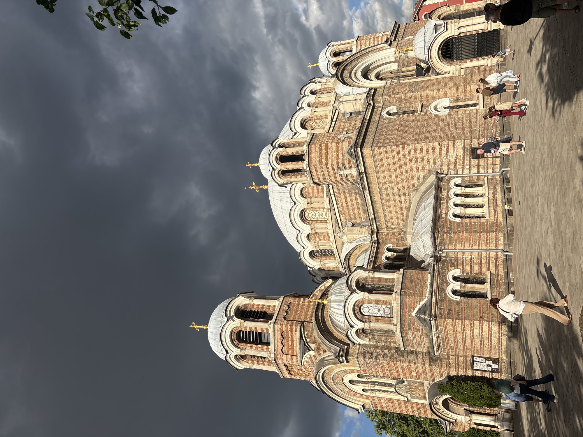 An exterior, low-angle, full shot captures the grandeur of an Orthodox church under a dark, dramatic sky. The church, constructed of reddish-brown brick with ornate architectural details, features multiple domes topped with golden crosses. The largest dome is centrally positioned, flanked by smaller domes and a prominent bell tower to the left. Arched windows with intricate detailing punctuate the facade. In front of the church, a paved plaza is populated by several individuals. A group of four women walks away from the camera towards the right side of the frame. To the left, two people, one appearing to be a child, walk away from the camera. Another individual walks towards the church entrance on the far right. The sunlight illuminates the front of the church while casting long shadows across the plaza. The sky is filled with heavy, dark clouds, creating a sense of impending weather.
