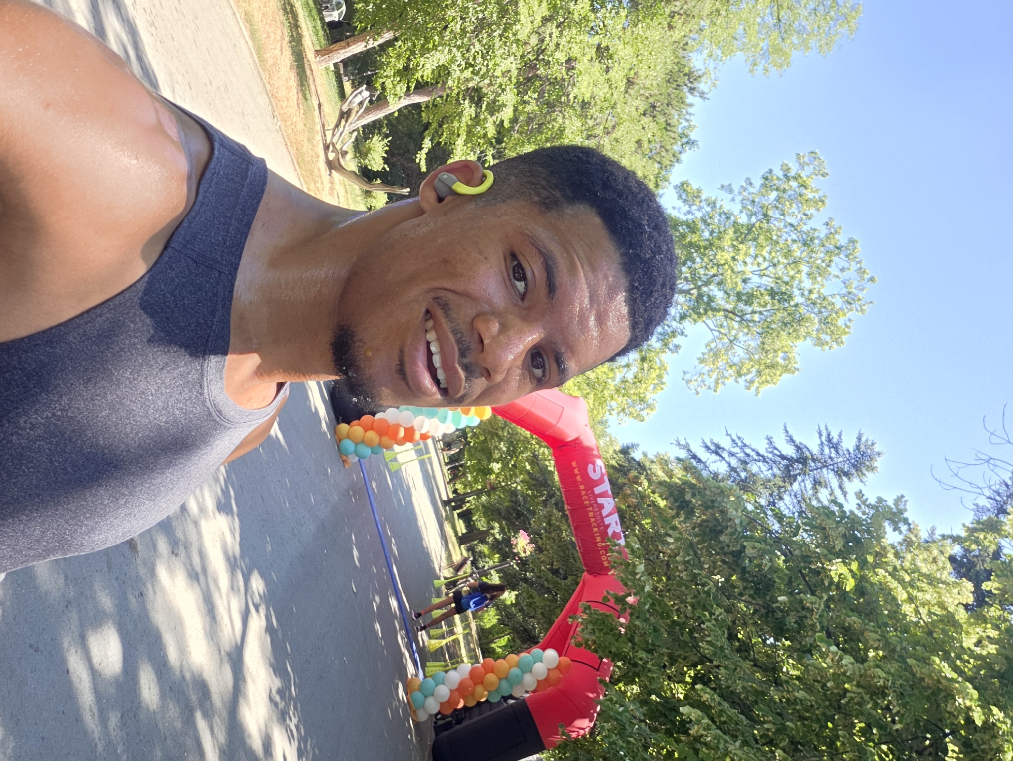 A man wearing a grey tank top and yellow earbuds is taking a selfie in a park on a sunny day. He appears to be sweating, suggesting he has been exercising. In the background, there is a red inflatable archway that says "START" and "WWW.RACETRACKING.COM", decorated with balloons. A woman with a backpack is visible behind the archway. The path in the park is lined with trees. The man's skin tone suggests he is of African descent. The scene appears to be at the beginning of a race or event.