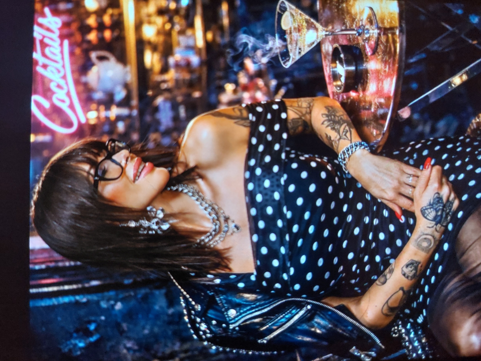 The image captures a woman in a vibrant, dimly lit bar or nightclub setting, likely located in Tsarevo, Bulgaria. She is shown from the chest up, positioned on her side and looking upwards with a wide, bright smile. She has dark hair with bangs and wears black-rimmed glasses, red lipstick, a prominent multi-strand necklace, and large statement earrings. Her attire consists of a black polka-dot dress layered with a black studded leather jacket.
Her arms are heavily tattooed with various designs, including a detailed butterfly on her right hand/wrist and other floral and abstract patterns on her forearms. She wears red nail polish, rings on her fingers, and a bracelet on her left wrist.
In the background, a glowing neon sign vertically spells out "Cocktails" in a cursive script on the left. Above her, a martini glass is featured, emitting wisps of smoke or vapor. The background is blurred with warm, ambient lights, contributing to a lively and nocturnal atmosphere. The woman appears happy and relaxed.