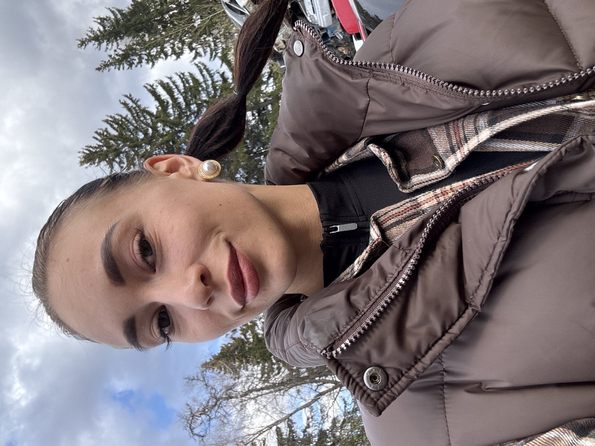 The image captures a close-up, low-angle perspective of a young woman outdoors in Chaikovsky, Russia. She is looking directly at the camera with a subtle smile, her head tilted slightly. Her dark hair is pulled back into a ponytail secured with a small elastic, and she wears a prominent gold-rimmed pearl earring on her visible ear. Her makeup includes defined eyebrows and dark eyelashes, and her lips are painted in a muted reddish-brown shade.
She is dressed for cold weather, wearing a brown puffer jacket with a metallic zipper and snap buttons visible. Underneath, a plaid shirt in shades of brown, white, and red can be seen, partially covered by the jacket. A black collared top with a short zipper closure is worn beneath the plaid shirt.
The background features a cloudy sky with patches of blue visible, suggesting an overcast day. Evergreen trees, likely spruce or fir, are prominent, along with bare deciduous trees, indicating either late autumn, winter, or early spring. Hints of what appears to be a vehicle with red and silver components are visible in the background, though indistinct. The overall scene suggests a cool or cold daytime environment. No discernible text is visible in the image.