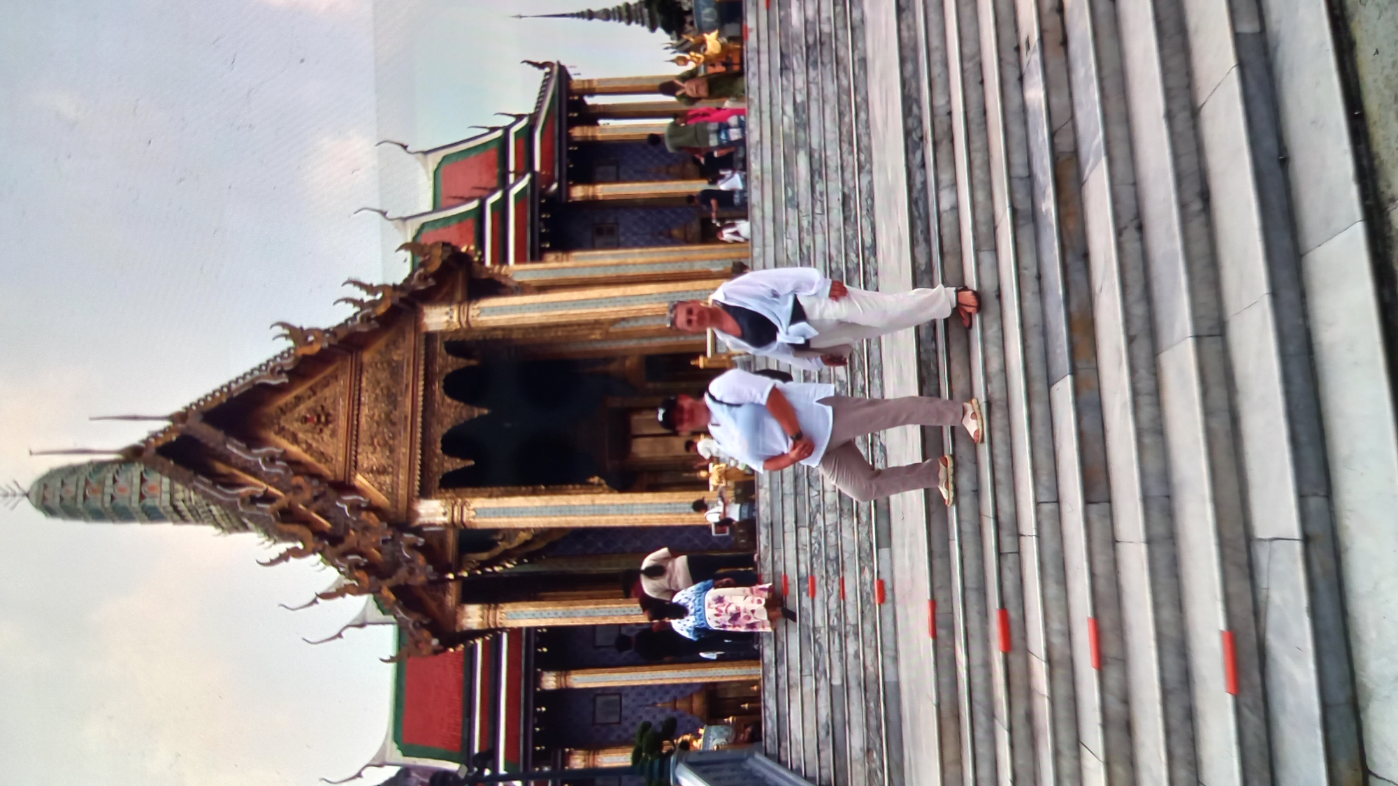 Two women are posing on a wide, marble staircase leading up to a temple structure. The woman on the left is wearing a white long-sleeved shirt, beige pants, and sandals. She has a black baseball cap on and a black backpack. The woman on the right is wearing a black tank top, a white unbuttoned shirt tied at the waist, and white pants. She is also wearing sandals. The temple behind them features ornate golden details, multiple tiered roofs with green and red accents, and tall, decorative pillars. Other people are visible in the background, some walking up the stairs and others standing near the temple entrance. The sky is overcast. The location is likely the Grand Palace in Bangkok, Thailand, not ตำบลเกาะพะงัน, which is an island.
