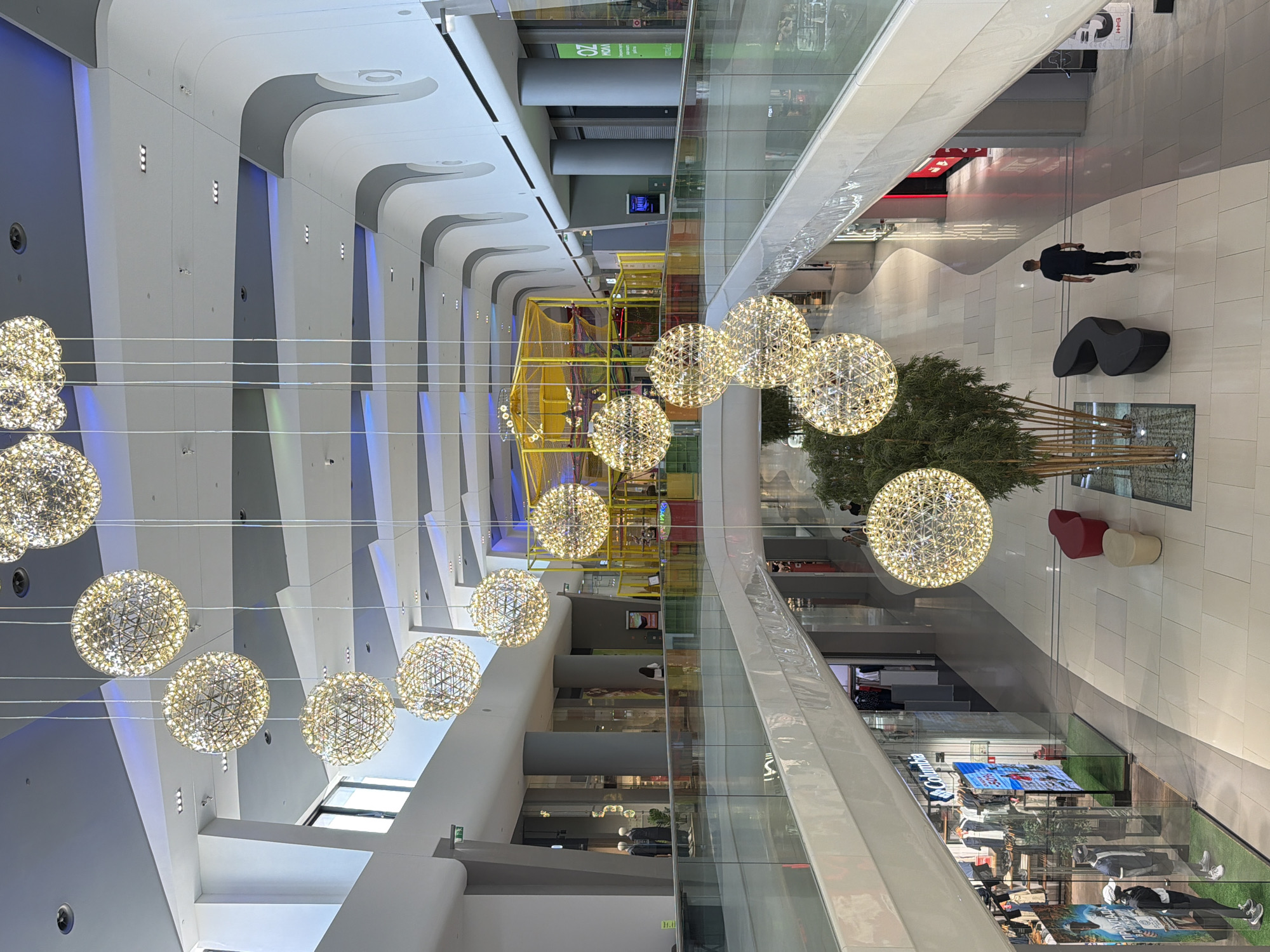 This is a photograph taken from an elevated vantage point within a modern shopping mall, likely in Krasnodar, Russia. The mall features multiple levels, with walkways and store fronts visible on each. The architecture is characterized by clean lines, white and gray tones, and a curved ceiling with integrated lighting, some of which is blue. Large, spherical chandeliers made of intricate, reflective material hang from the ceiling, illuminating the interior.
In the foreground, a glass-paneled walkway overlooks the main floor of the mall. Below, on the ground level, a man in a dark t-shirt and black pants walks away from the camera towards the right. Further to the left, on the ground floor, are several shops with large glass windows displaying merchandise and mannequins. One shop appears to be "Columbia." Nearby, there are minimalist benches and some decorative bamboo-like plants.
On an upper level, a brightly colored indoor playground with yellow netting is visible. Various shoppers are sparsely distributed throughout the mall, some on the walkways and others on the ground floor. The overall impression is one of a spacious, well-lit, and contemporary commercial environment.