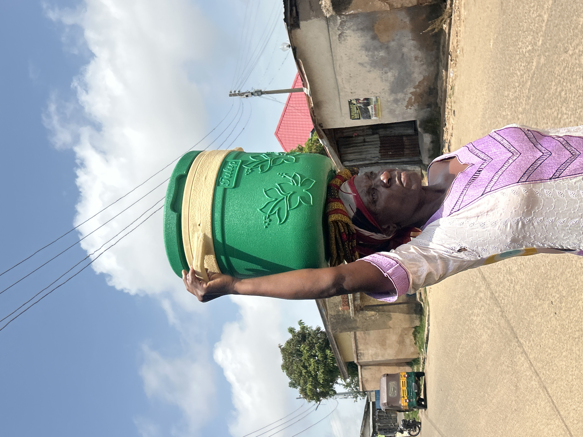 A woman walks along a dusty road in the city of Jos, Nigeria, carrying a large green water container balanced on her head. She wears a white and purple patterned dress. The background shows buildings with corrugated metal and stucco walls, and a red-roofed structure. A small three-wheeled vehicle and a motorcycle are visible on the road. Power lines stretch across the sky, which is partly cloudy. The image is taken from a medium close-up perspective, focusing on the woman and the water container.