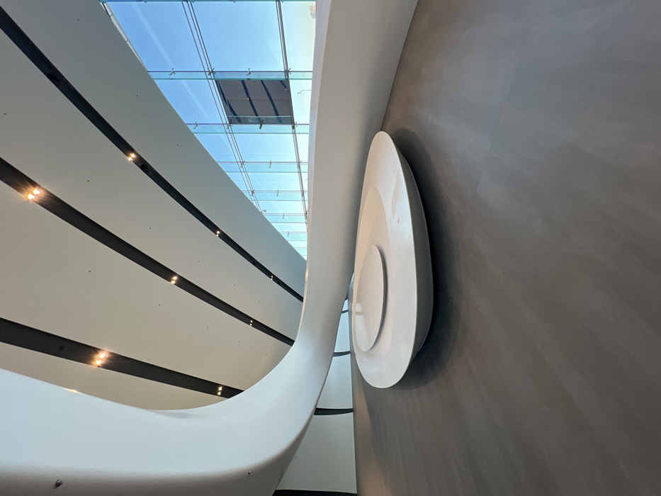 The image depicts a modern, minimalist interior space, identified as part of the Lakhta Center in Saint Petersburg, Russia. The scene is characterized by smooth, curvilinear white architectural elements set against a dark grey floor and a large glass curtain wall. A prominent, sweeping white structure originates from the upper left, curves downwards, and extends horizontally towards the right. The white ceiling features parallel diagonal dark strips containing evenly spaced, warm-toned recessed downlights. The entire right side of the frame is a multi-paneled glass wall, revealing a clear blue sky and distant horizon outside. In the lower-center, on the dark grey floor, rests a low, asymmetrical white sculptural object with an elevated circular surface, resembling modern furniture or a display piece. The area is devoid of people, objects, actions, or interactions, presenting a pristine, brightly lit, and spacious contemporary environment.