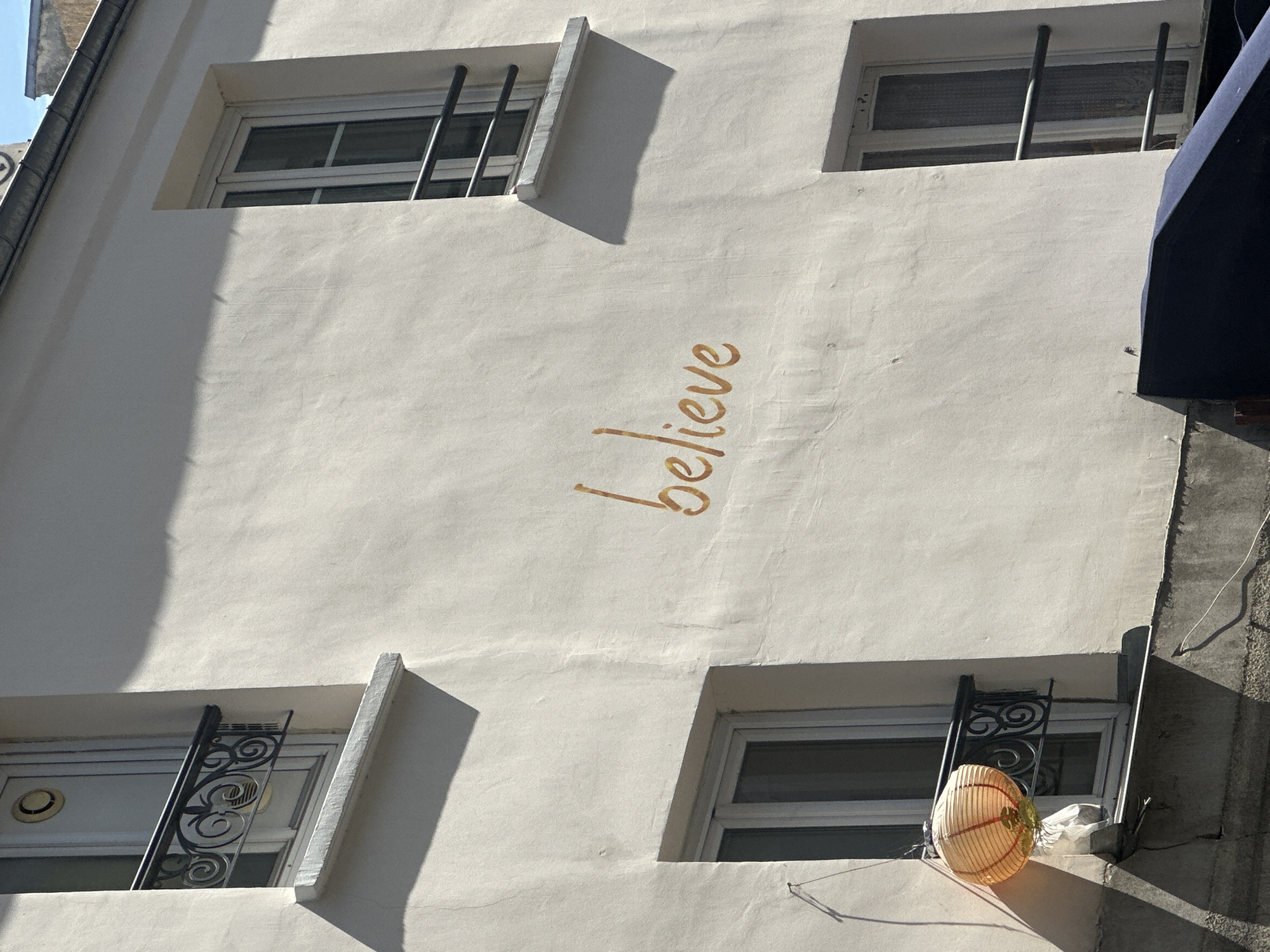 A low-angle, sunlit view of a cream-colored building facade in Paris, France. The word "believe" is written vertically in a brown script on the wall. Four windows are visible, with security bars or decorative wrought iron on three. A Chinese lantern rests on the windowsill of the lower right window. A section of a dark blue awning is visible at the upper right corner. The building exterior appears weathered.