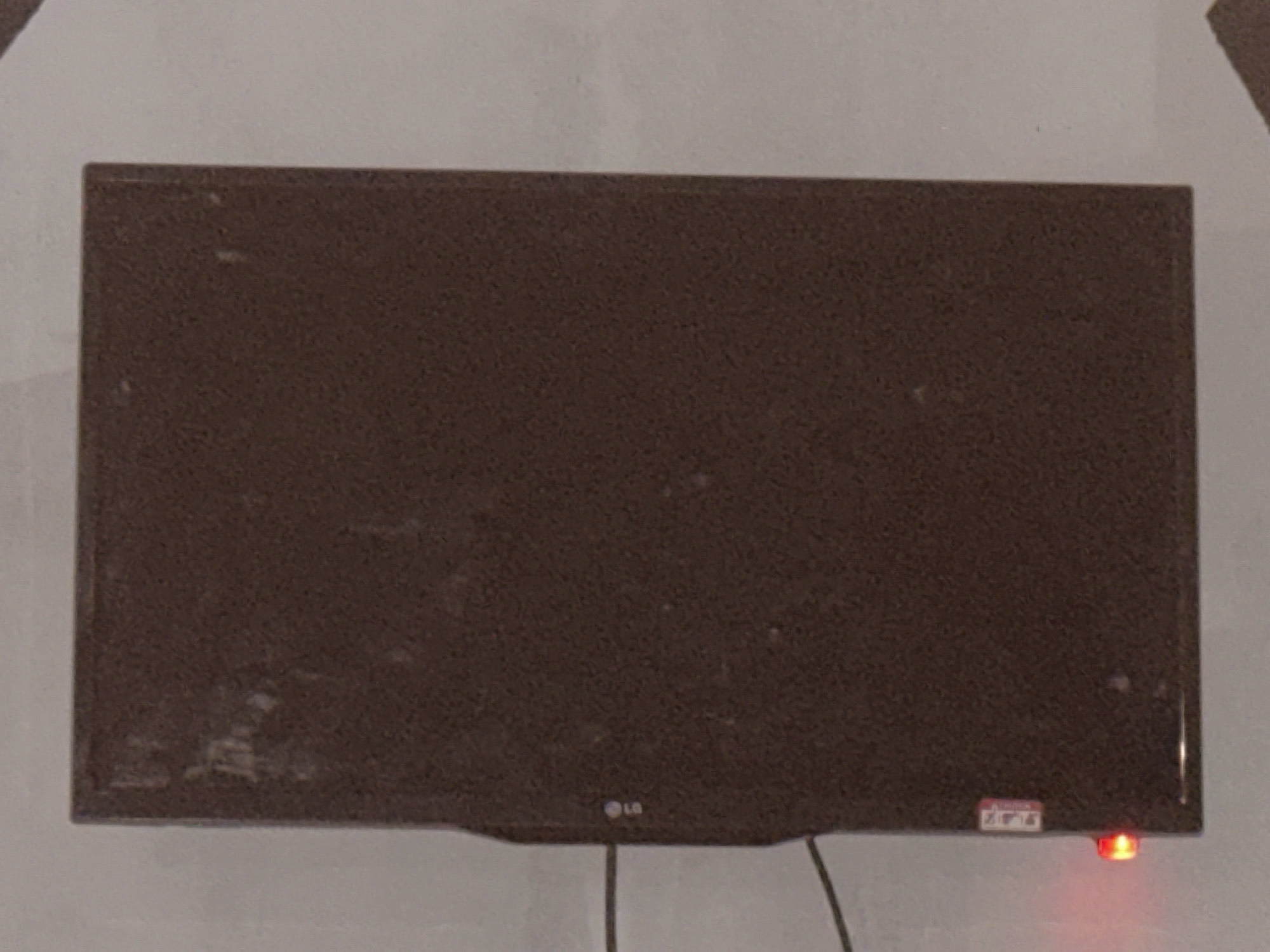 The image shows a dark brown LG television mounted on a wall. The screen is dark, and there is a small red indicator light on the lower right side of the television. There is also a small white sticker with some red text and symbols on the lower right bezel of the television. The wall appears to be painted a light color, possibly white or off-white. There are some smudges and marks on the television screen and bezel. The image quality is grainy, suggesting it was taken in low light conditions. There is no indication of the location in Jos, Nigeria.