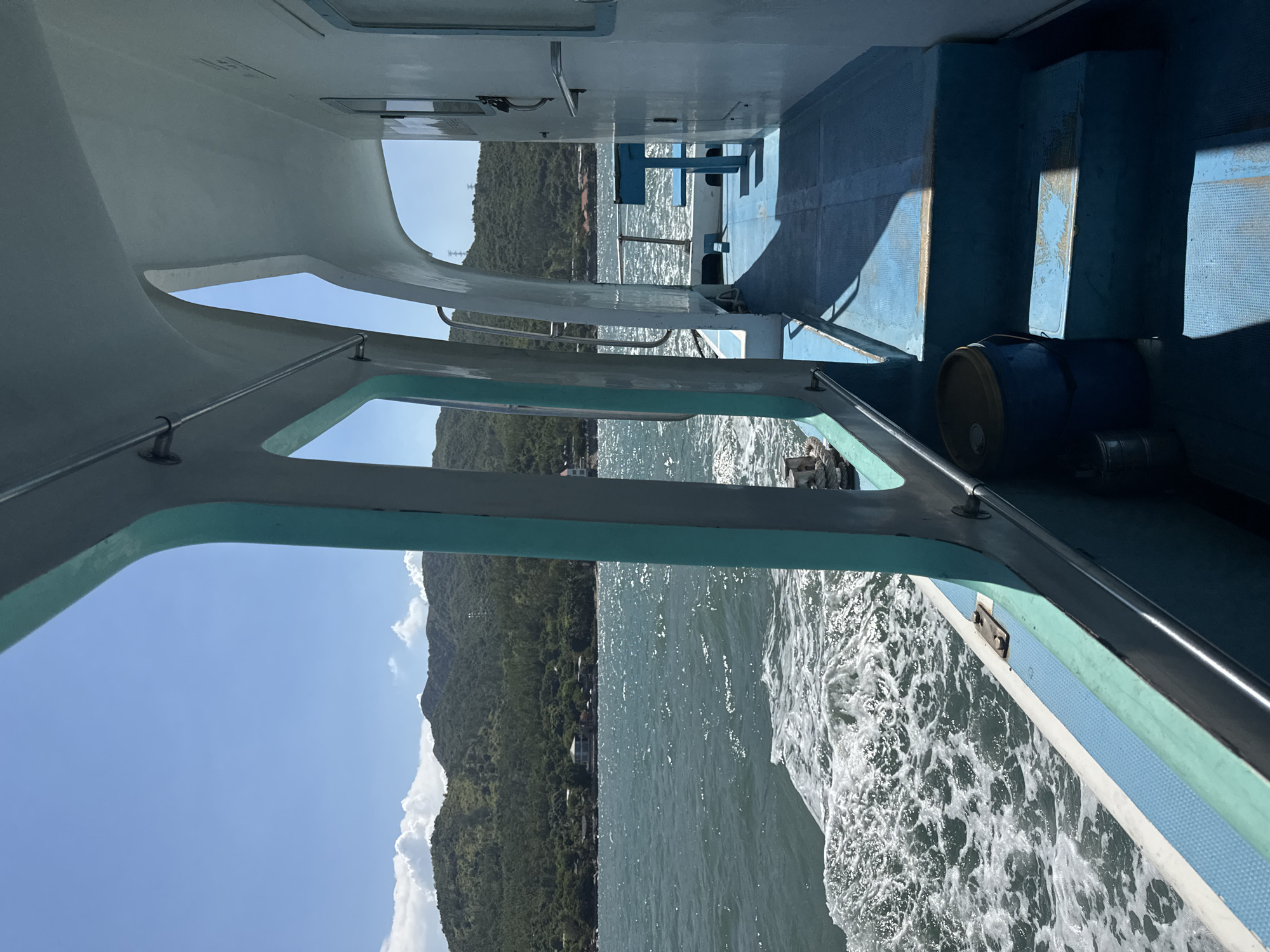 The image shows the interior of a boat moving on the water, with a view of a coastline and mountains in the distance. The boat's deck is blue with some white and turquoise trim. There are steps leading up to a higher level of the deck, where a blue bench and table are visible. A blue bucket and some smaller containers are on the lower deck. The water is choppy, with white foam trailing behind the boat. The coastline is lined with green trees and some buildings, and the mountains behind it are also covered in vegetation. The sky is clear blue with some white clouds. The scene suggests a sunny day and a leisurely boat trip.