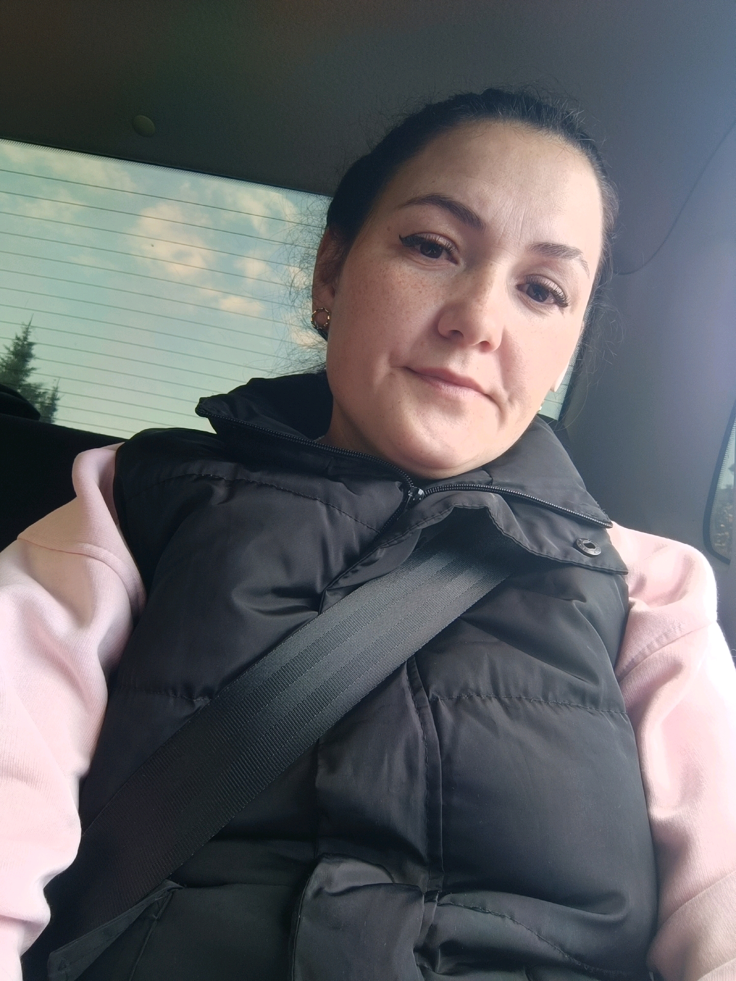 In a close-up selfie taken from the passenger seat of a car, a woman with fair skin and freckles is captured. She is looking directly at the camera with a neutral expression. Her dark hair is pulled back, and she is wearing a black puffer vest over a light pink hoodie. A black seatbelt is diagonally across her chest, securing her. The background reveals the rear window of the car, through which a cloudy sky and a blurred green tree are visible. The image does not provide any visual cues to determine the specific location within the city of Perm, Russia.