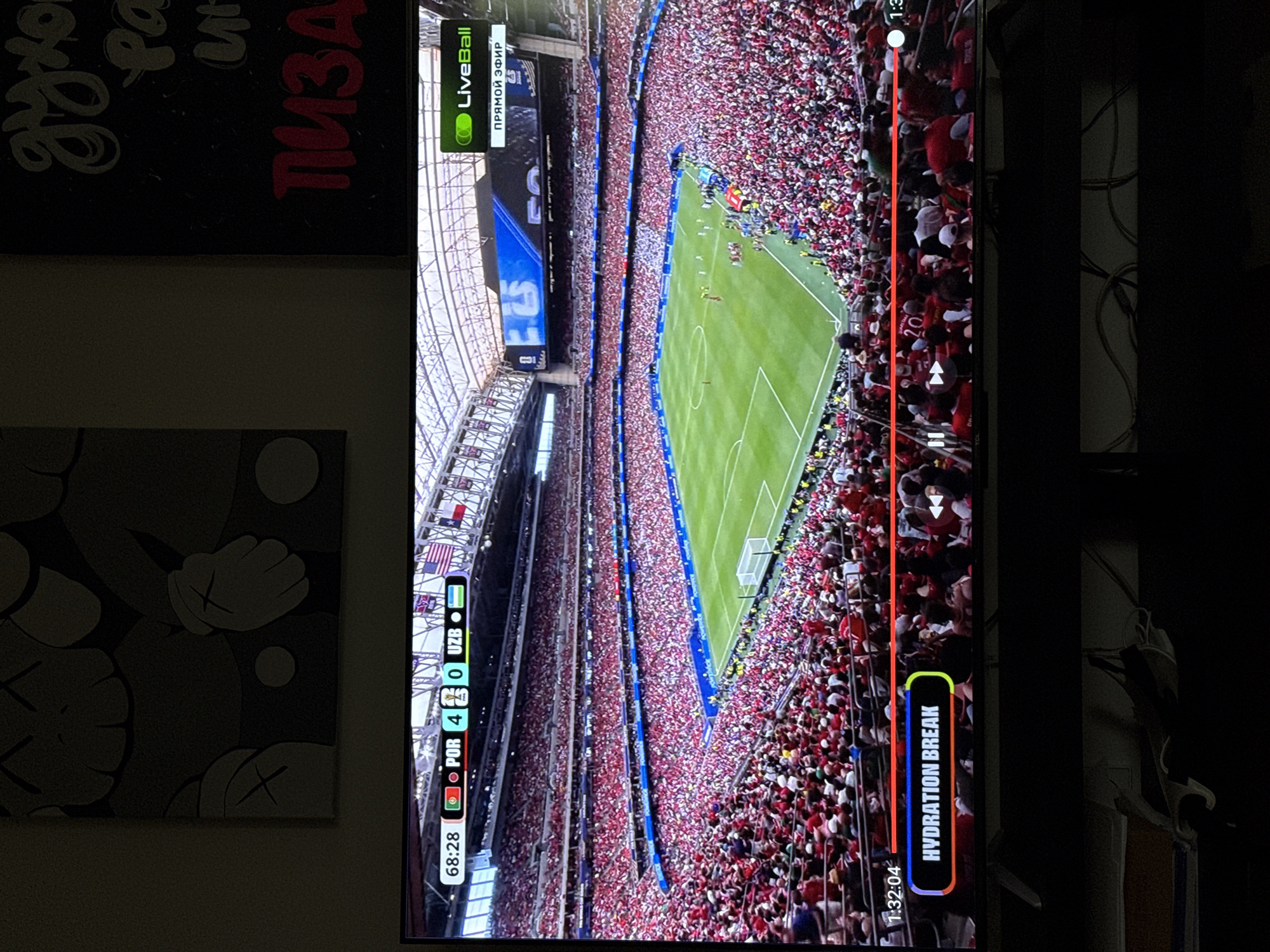 This is a vertical, eye-level medium shot of a soccer match being played in a packed stadium. The focus is on the field and the expansive crowd. The field is a vibrant green, marked with white lines, and a goal is visible in the distance. Spectators are packed into the stands, mostly wearing red clothing, creating a sea of color. The overall impression is one of intense energy and a large gathering of people.
In the foreground, on the left side of the image, there's a section of a TV screen displaying a portion of the game. This overlay includes game information such as "68:28" (likely indicating the current game time), team scores "POR 4" and "UZB 0", and a "LiveBall" logo with "ПРЯМОЙ ЭФИР" (meaning "Live Broadcast" in Russian). On the right side of the screen, a "HYDRATION BREAK" notification is visible, along with a timestamp "1:32:04".
The lighting appears to be artificial, typical of an indoor stadium setting or a broadcast of a nighttime game. The background is dark, suggesting the surrounding area outside the stadium is unlit or obscured. The image is framed such that it captures a significant portion of the stadium's capacity, emphasizing the scale of the event. The presence of the overlaid graphics on the screen indicates this is a media capture of a broadcast, not a direct view of the event itself.