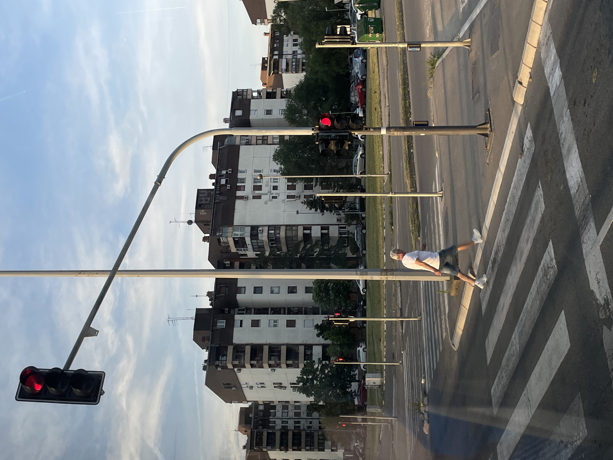 A pedestrian is crossing a zebra crossing in Belgrade, Serbia. The man is wearing a white t-shirt, shorts, sneakers, and a baseball cap. Traffic lights are red. Tall apartment buildings are located in the background.