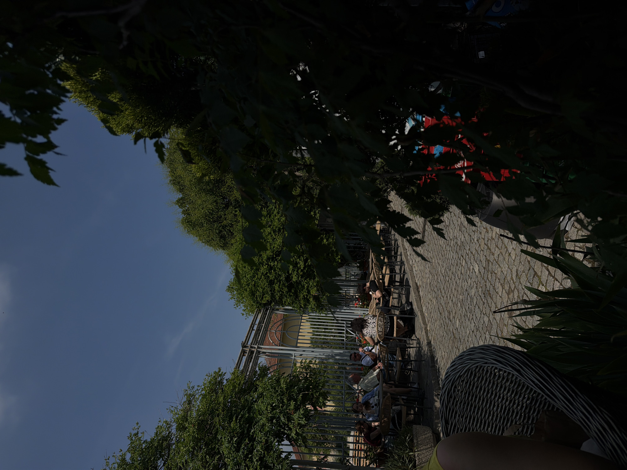 The image captures a lively outdoor cafe or restaurant patio on a bright, sunny day, likely in Plovdiv, Bulgaria. The scene is viewed from a low perspective, as if from a seated position, with part of a dark grey wicker chair and a person's arm visible in the bottom right foreground.
The patio features cobblestone pavement, on which rows of tables and chairs are arranged. Several patrons are seated, enjoying the weather and each other's company. People are seen conversing and relaxing. Notable individuals include a person wearing a distinctive leopard print top and a man in a white shirt and sunglasses.
To the left of the patio, lush green trees and dense foliage create a natural canopy, casting shadows across parts of the scene. A tall metal fence or trellis structure runs vertically through the middle ground, separating the cafe area from what appears to be a street or pathway beyond.
In the background, a portion of a building with a yellowish facade and a reddish roof is visible through the gaps in the greenery. Stacked beverage crates, some red and some blue, are discernible in the distance. While not perfectly clear, a white logo resembling "Coca-Cola" can be faintly seen on one of the red crates.
The clear blue sky, bright sunlight, and long shadows indicate midday or afternoon. The overall atmosphere is relaxed and social, typical of a European cafe setting. No explicit text other than the faintly visible potential "Coca-Cola" logo is clearly identifiable.