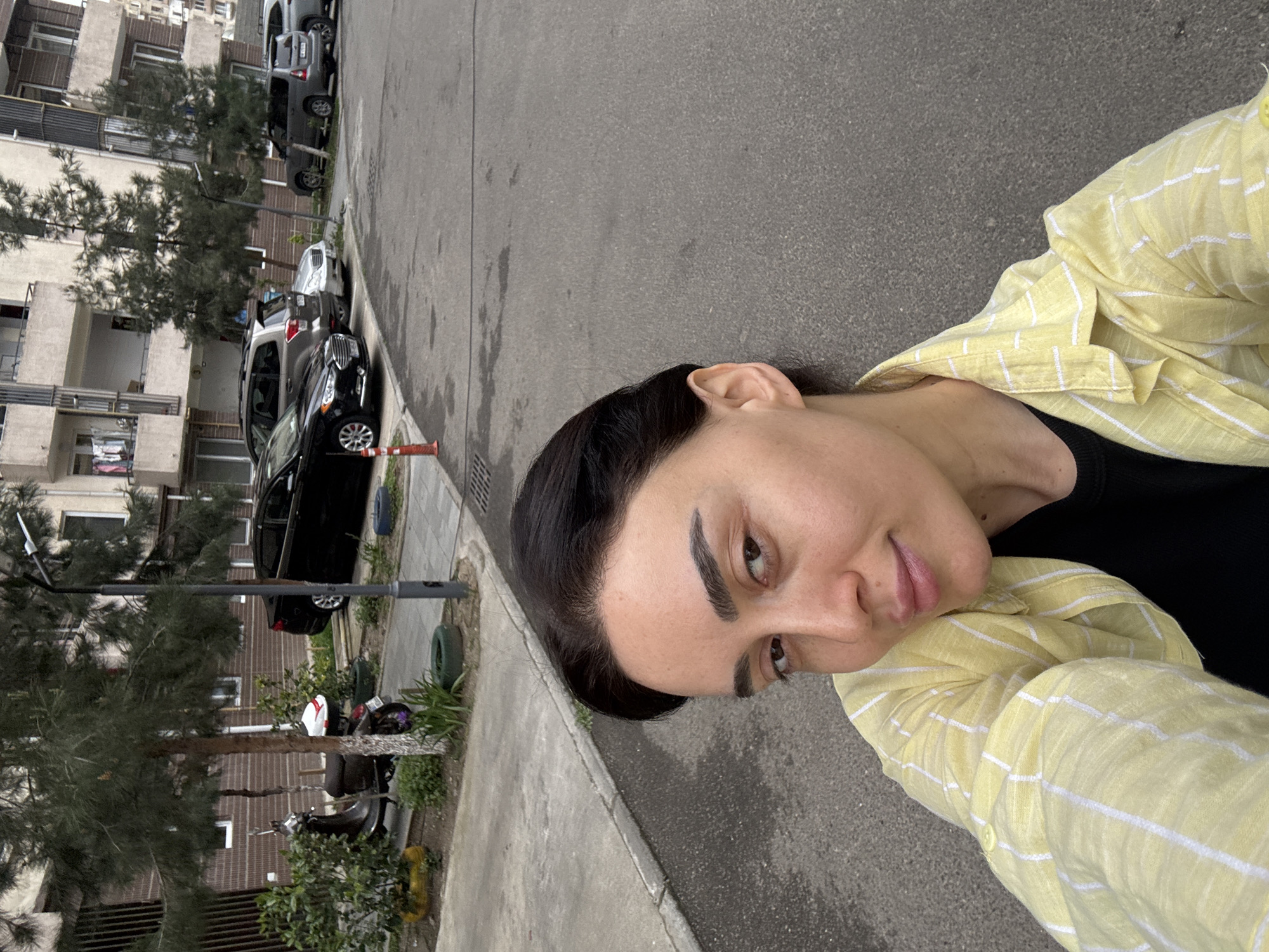 A woman takes a selfie outdoors, positioned in the right foreground of the vertically oriented image. She has dark hair, dark eyebrows, and is wearing a black t-shirt under a light yellow, white-striped collared shirt. Her expression is neutral as she looks directly at the camera.
The background depicts an urban street scene in what is identified as Tbilisi, Georgia. An asphalt road occupies the middle ground, with a concrete sidewalk and curb running along its left edge. Several vehicles are parked along the sidewalk, including a black sedan in the mid-ground, followed by a silver SUV and a grey SUV further back. A smaller grey car is also visible in the distance. An orange traffic cone is partially visible near the black sedan.
Behind the parked cars and sidewalk are multi-story apartment buildings with light-colored facades and balconies. Pine-like trees are scattered between the road and the buildings, providing some greenery. A street lamp pole is also visible. Further down the street, on the sidewalk, a scooter or moped is parked near some low bushes. The overall lighting suggests it is daytime, likely a clear or partly cloudy day, with no harsh shadows. There is no visible text in the image.