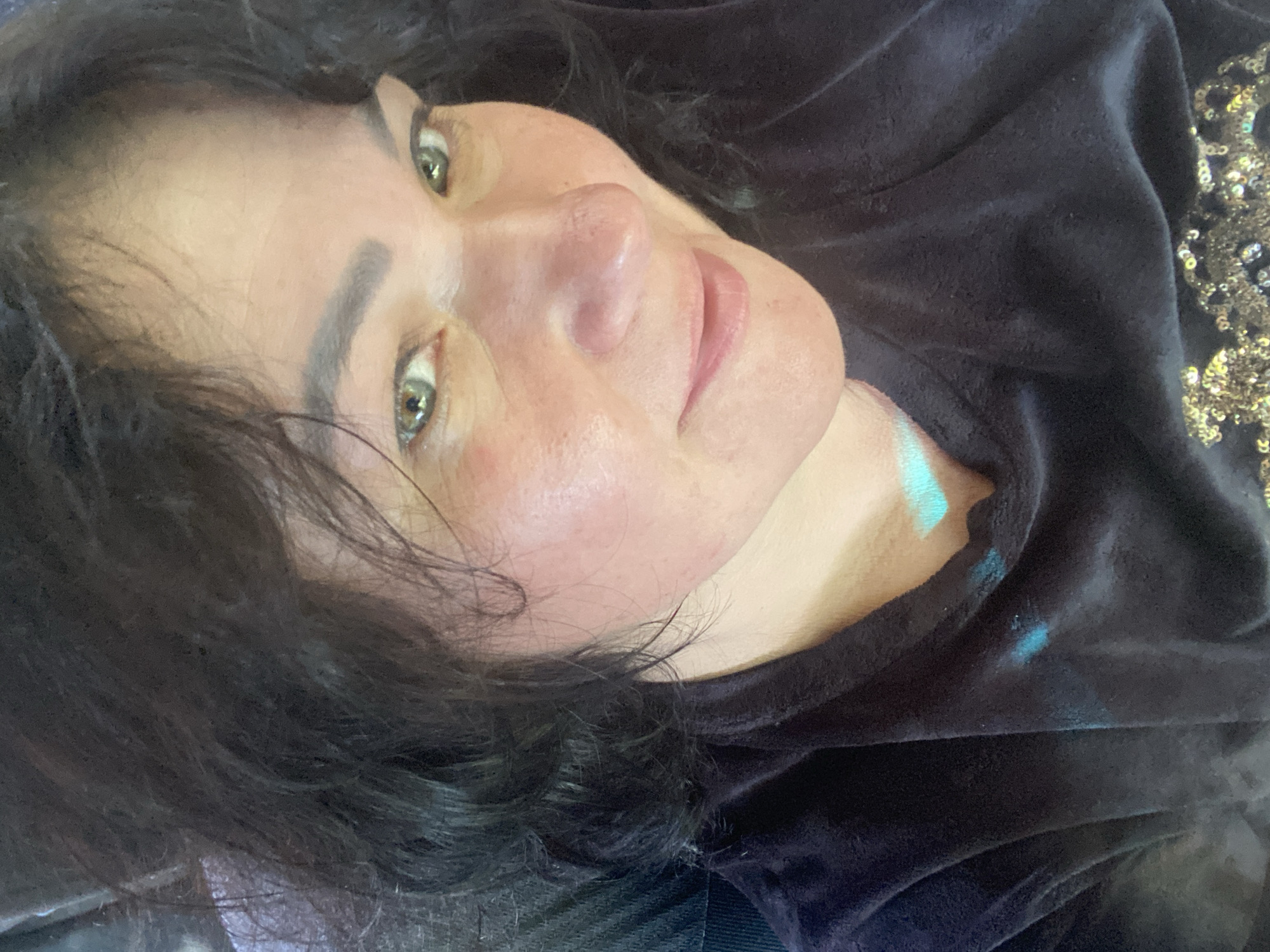 A close-up selfie of a woman with dark hair and green eyes, facing the camera. She is wearing a dark-colored, velvety shirt with gold sequins on the lower part of the shirt. The image appears to be taken indoors, possibly in a home environment. There are no identifiable landmarks or clues to indicate the specific location within the city of Dnipro, Ukraine. The lighting suggests an artificial light source.