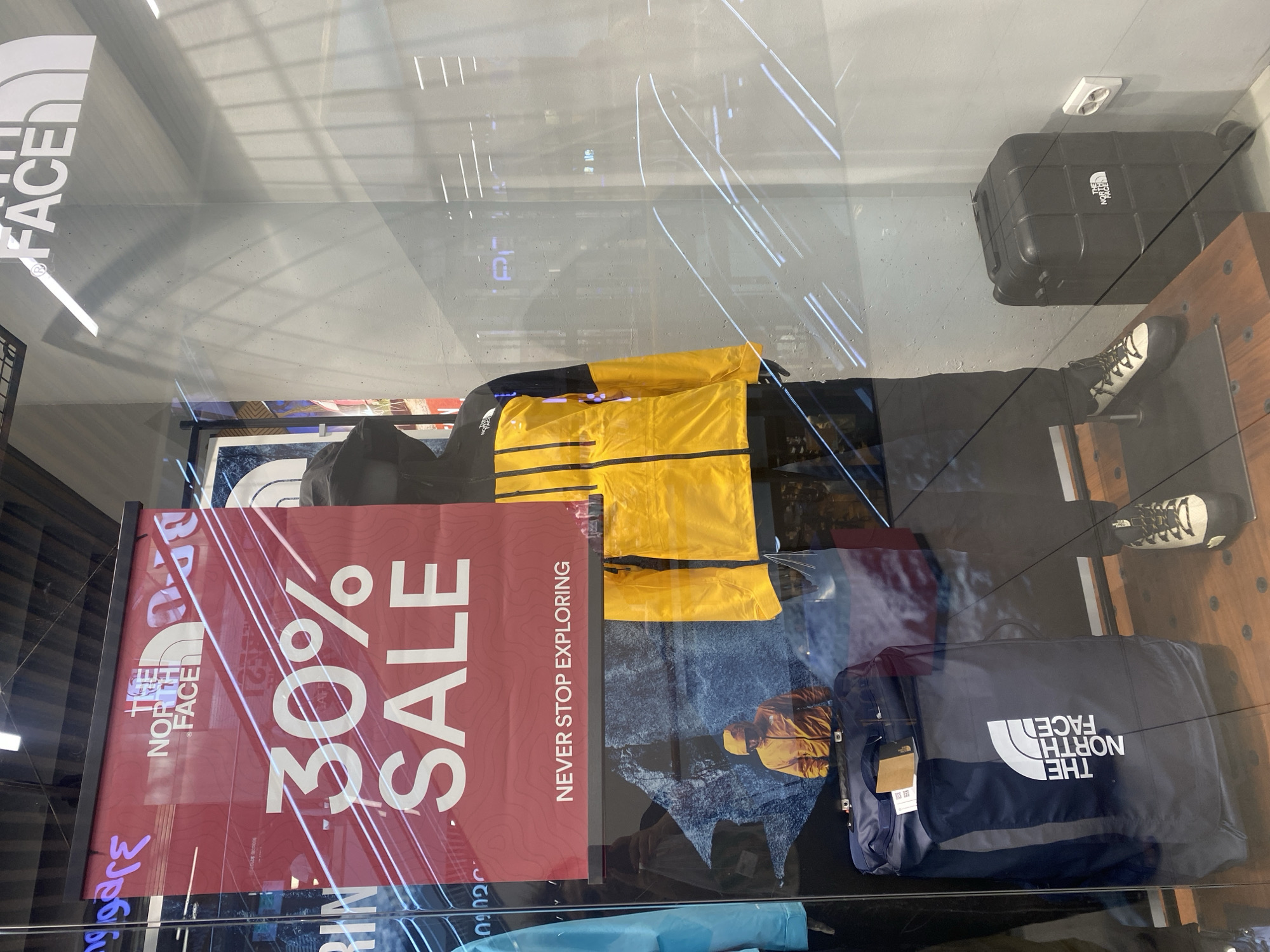 A window display for "The North Face" is shown, featuring a large red banner with "30% SALE" and "NEVER STOP EXPLORING" printed on it. A mannequin is dressed in a yellow and black jacket and black pants. To the left of the mannequin is a dark blue duffel bag with "The North Face" logo. To the right is a small black suitcase with wheels and the same logo. In the background of the display, a man wearing a yellow jacket is depicted in a mountainous landscape. The reflection of lights and interior of a store are visible on the glass. The location is identified as The North Face store in Tbilisi, Georgia.