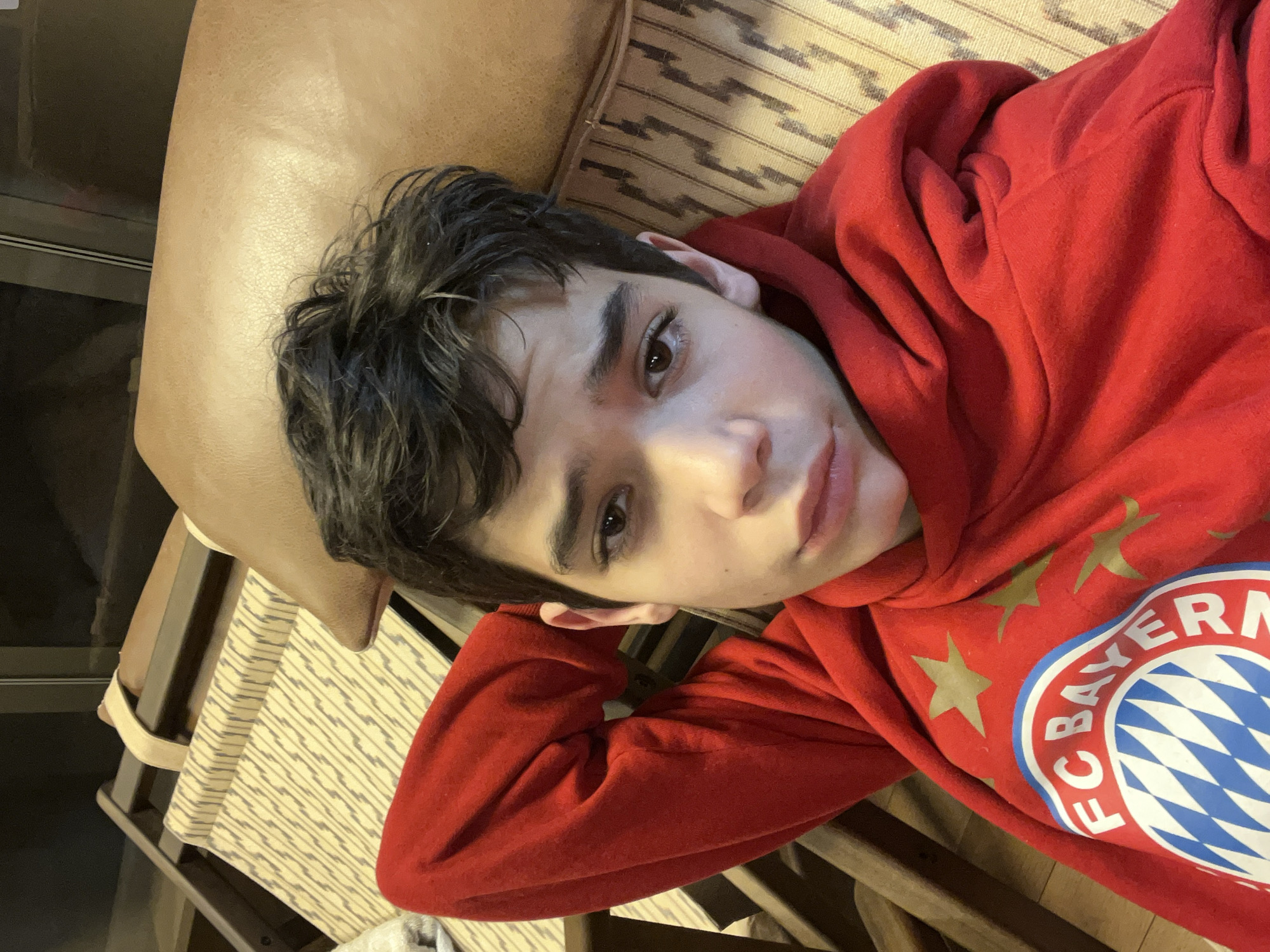 A young person with dark hair is reclining on a chair or sofa with a brown leather-like cushion. They are wearing a red hooded sweatshirt with the FC Bayern Munich logo on it. The seating appears to be made of wood with a woven fabric covering. The background is blurry and dark. There is no indication of this picture's location being in Sofia, Bulgaria.