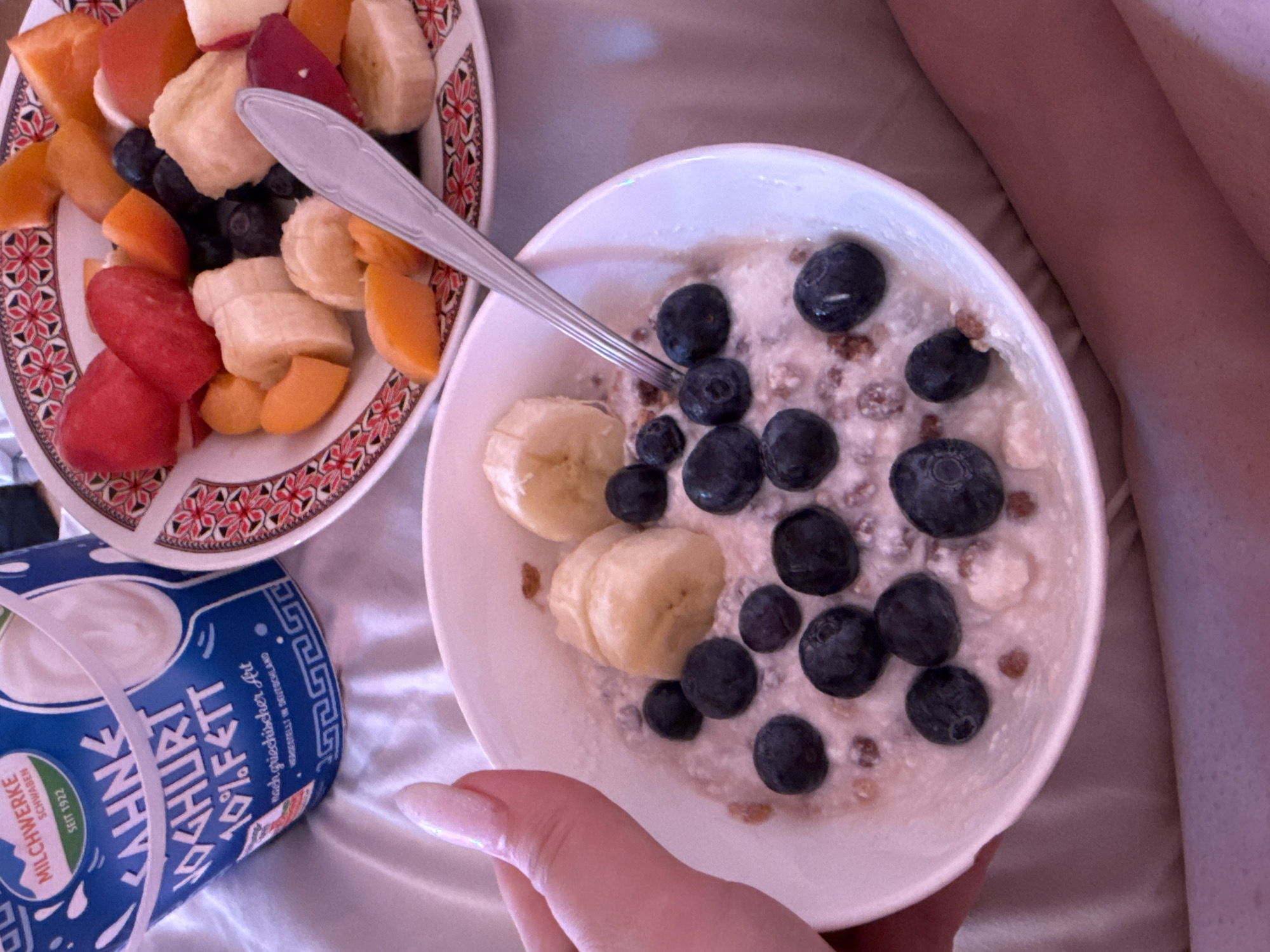 The image captures a close-up, elevated view of a person enjoying a healthy meal or snack in what appears to be a relaxed, indoor setting. The primary focus is on two dishes of fruit and a container of yogurt.
A white ceramic bowl, held by a hand with light-colored, possibly manicured nails, contains a creamy mixture, likely yogurt or oatmeal, topped with numerous whole blueberries, several sliced bananas, and scattered granola or cereal pieces. A silver-colored spoon rests partially in this bowl.
To the left of this, on a decorative white plate with intricate red and black geometric patterns, is an assortment of cut fresh fruit. This plate includes vibrant red pieces (possibly strawberries or plums), bright orange slices (suggesting apricots or melon), yellow banana chunks, and some additional blueberries.
In the bottom left corner, a blue and white yogurt container is prominently displayed. The text on the container clearly reads "ZAHNE YOGHURT 1.5% FETT" (1.5% fat), "MILCHWERKE SCHWABEN SEIT 1922" (Dairy Works Swabia Since 1922), and "Hergestellt in Deutschland" (Made in Germany), indicating its origin. The container also features a Greek key pattern along its top edge.
The background consists of light-colored, possibly satin or silk, fabric, suggesting a bed or sofa. A portion of a person's upper leg or thigh, covered in a peachy-beige fabric, is visible in the top right, reinforcing the casual, relaxed atmosphere. The overall scene conveys a moment of comfortable, healthy indulgence, possibly a breakfast or snack time.