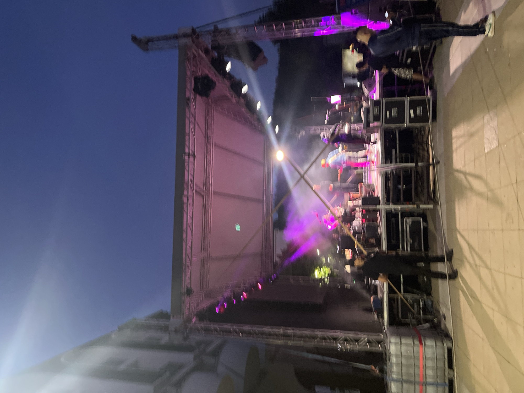 The image captures an outdoor evening event at an unknown city location. A brightly lit stage with pink and purple lighting is set up, featuring a band performing with instruments and microphones. Several individuals, likely musicians, are on stage, with one person holding a guitar. Stage equipment, including speakers and lighting rigs, is visible. In the foreground, a man in black clothing is holding a long pole, possibly a crew member. Other spectators are also present, some in the background near the stage and others in the lower right corner of the image. The environment appears to be a public space, with a paved ground and a building facade to the left, suggesting an urban setting. The sky is a deep blue, indicating dusk or early night.