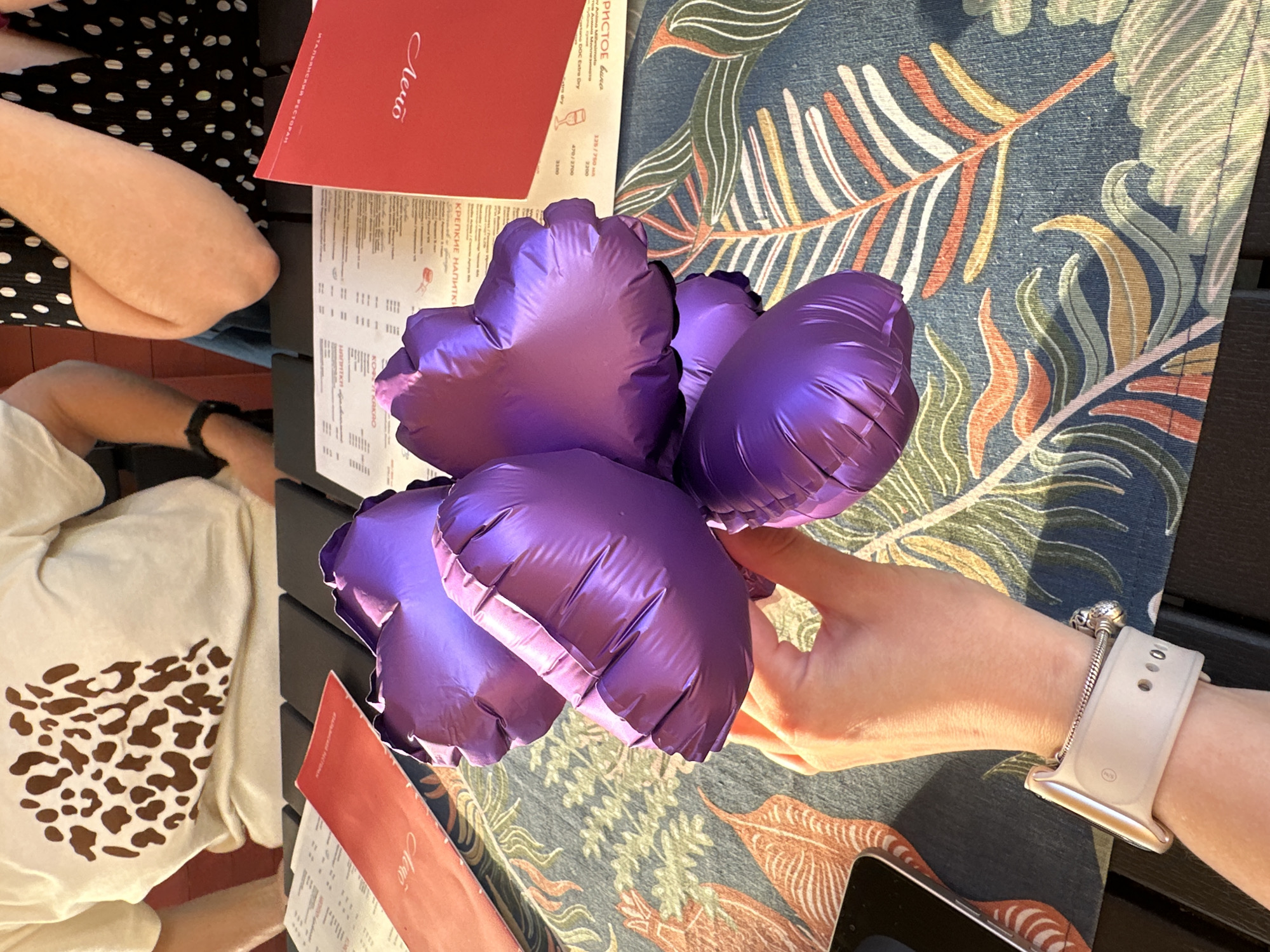 The image, likely taken in Perm, Russia, shows a table setting at what appears to be a restaurant called "Лечо" (Lecho). A person is holding a bouquet of small, purple heart-shaped balloons. On the table, there's a red menu with the restaurant's name, a menu card listing prices in Russian rubles, and a table covering with a tropical leaf pattern. A person is wearing a white shirt with a leopard print and another person is wearing a black and white polka dot dress. The person holding the balloons has a white watch on their wrist. A smartphone is partially visible on the table. The lighting suggests an outdoor or well-lit indoor setting.