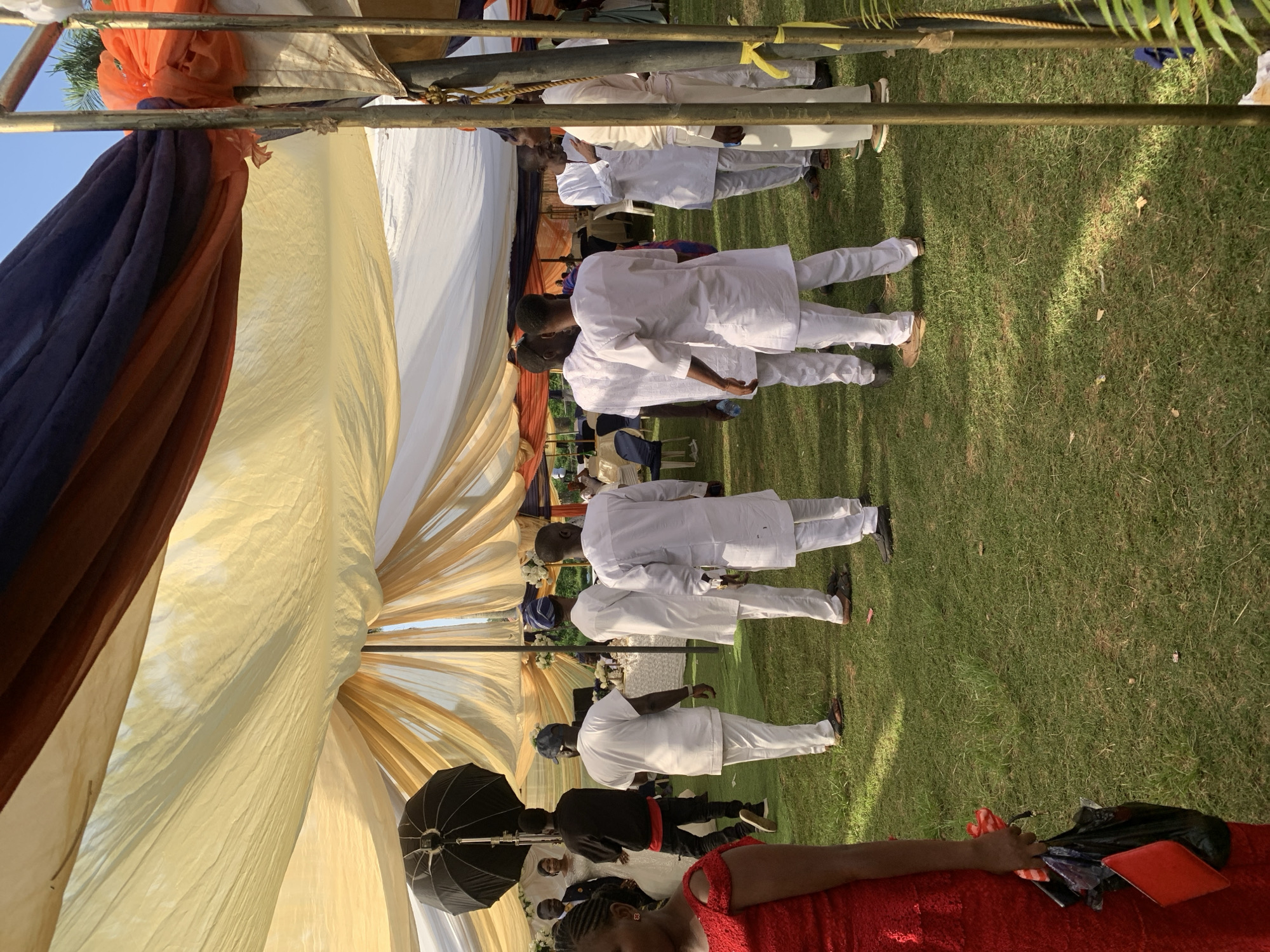 The image shows an outdoor event, possibly a wedding or a celebration, occurring on a grassy lawn. Several people dressed in white attire are standing and walking in rows, suggesting a procession or a formal entrance. A tent or canopy is set up above the gathering, with fabric drapes in shades of white, gold, orange, and dark blue or purple. Other details include an umbrella with soft light.
The image's location is described as Jos, Nigeria, but this cannot be determined from visual elements alone.