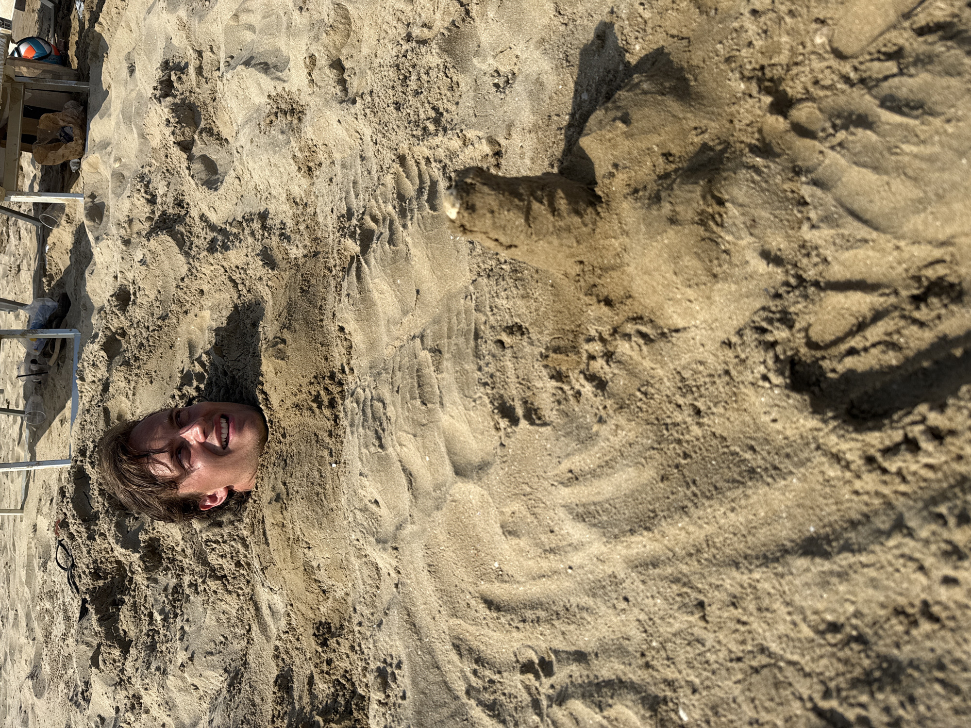 A man is buried in sand on a beach, with only his head and shoulders visible. He is smiling and looking to his right. In the foreground, a small sandcastle is being built. The location is the beach in Rimini, Italy, suggested by the presence of a beach umbrella in the background and the overall setting. The sun is shining, casting shadows on the sand.