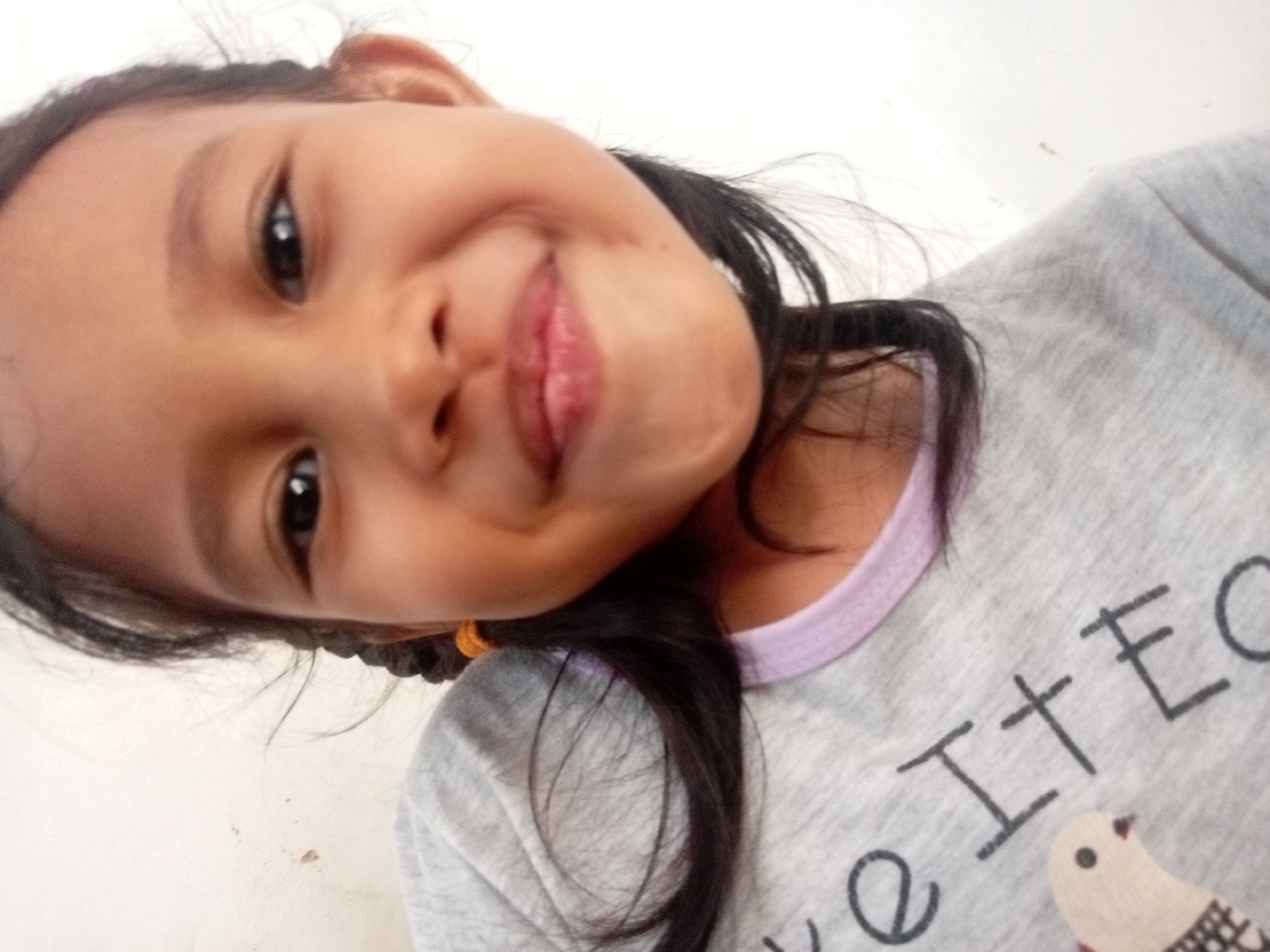 The media file is a close-up, slightly angled, portrait-style photograph of a young girl. She appears to be of Asian descent and is looking directly at the camera with a playful, slightly mischievous expression, her mouth open in a smile. Her dark hair is partially visible, with a few strands falling across her face. She is wearing a grey t-shirt with a light purple trim around the neckline. There is visible text on her t-shirt, partially obscured, which appears to read "e It E C" and a graphic of a small bird. The background is a plain, light-colored wall, suggesting an indoor setting. The lighting is bright, indicating it could be daytime. The subject's youthful features and cheerful demeanor suggest a casual, everyday moment. The overall impression is one of a happy and engaging child. The location context is Batam, Indonesia.