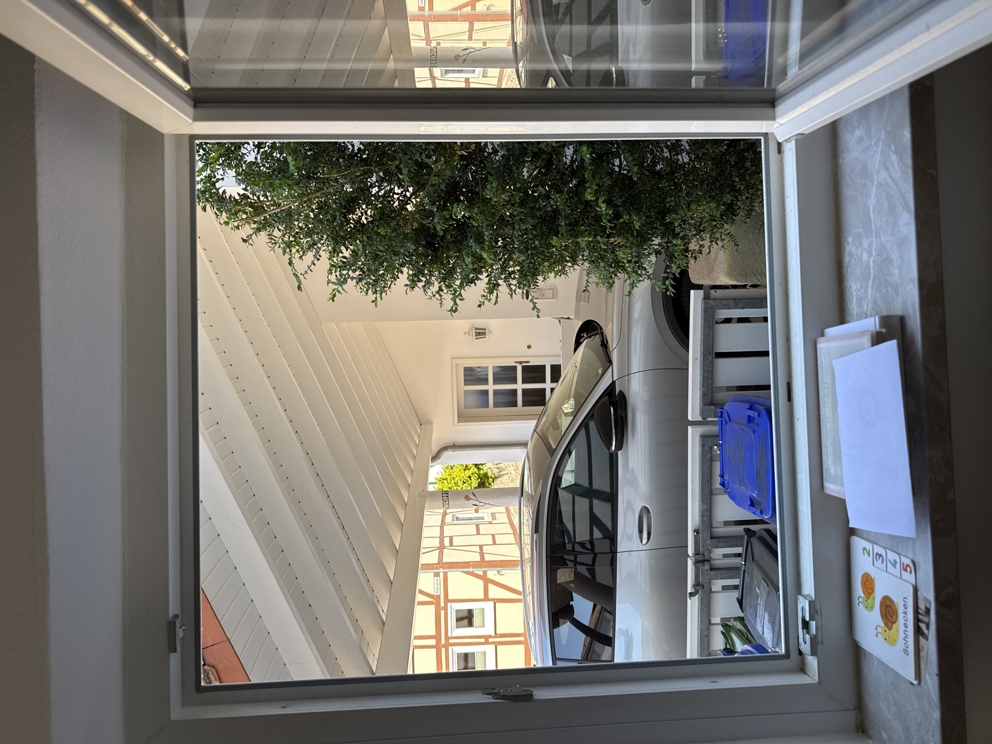 The image is an interior shot looking out of a window at a covered outdoor space. Through the window, a white car is parked under a sloped roof structure with white beams. Sunlight is illuminating the scene, suggesting it is daytime, and the weather appears clear.
In the foreground, the window frame is visible, and to the right, a tiled surface with some papers and a children's card featuring snails and numbers are placed. The card has the word "Schnecken" printed on it.
The background shows a traditional building with exposed timber framing and yellow walls, indicating a European architectural style. Green foliage from a tree is also visible, framing the view of the car and the building. The scene appears to be a residential or commercial area, possibly a driveway or carport next to a house. No people are visible in the image. The overall mood is calm and domestic.