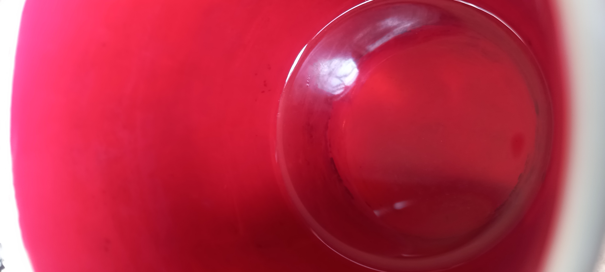 A top-down, close-up shot of a red liquid contained within a white receptacle. The liquid fills the frame, with a round area on the right side showing more depth, as if in a curved container. Reflective highlights appear on the liquid's surface. No people or identifiable objects are present other than the liquid and container. The location is not discernible from the image.