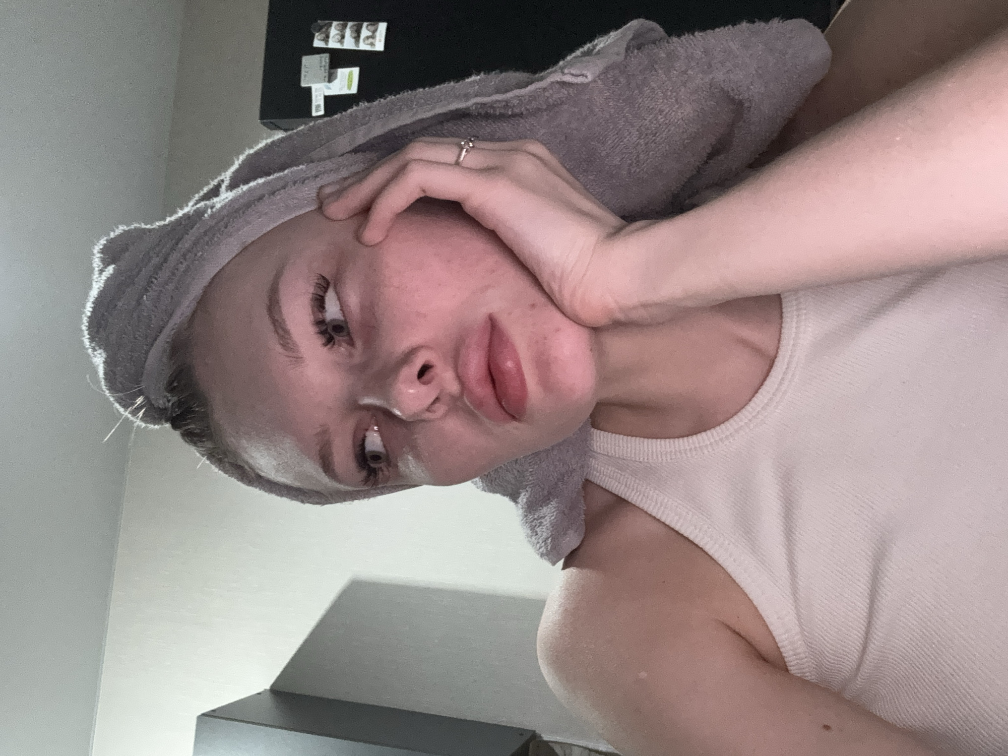 A person with fair skin and a towel wrapped around their head is visible. The person is wearing a white tank top. The person's left hand is touching their face. The background includes a wall, cabinets, and a refrigerator with magnets on it. The image shows an indoor location, likely a bathroom or bedroom. The location in the city of Perm, Russia is not discernible from the image.