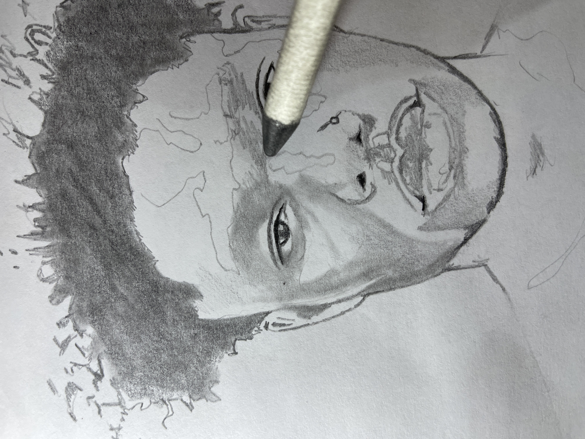 The image displays a close-up of a grayscale pencil drawing of a person's head on white paper, seen from the upper chest up. The drawing depicts a person, likely male, with short, dark, textured hair that appears to be in progress, with dense shading applied. The facial features are partially rendered: the left eye is shaded, with clear light reflections, and the left side of the face shows contrast and shading. The nose is outlined and lightly shaded, featuring a small nose ring on the right nostril. The mouth is open, revealing teeth, and shaded to create depth. The right eye and forehead are mostly outlined with light pencil strokes, indicating an unfinished state. A pencil blender or tortillon, with a light-colored body and dark tip, is positioned diagonally on the right side of the image, its tip touching an area near the right eyebrow and temple of the drawn figure, suggesting an active blending process. The background is the plain white texture of the paper, providing no environmental context beyond the drawing surface.
