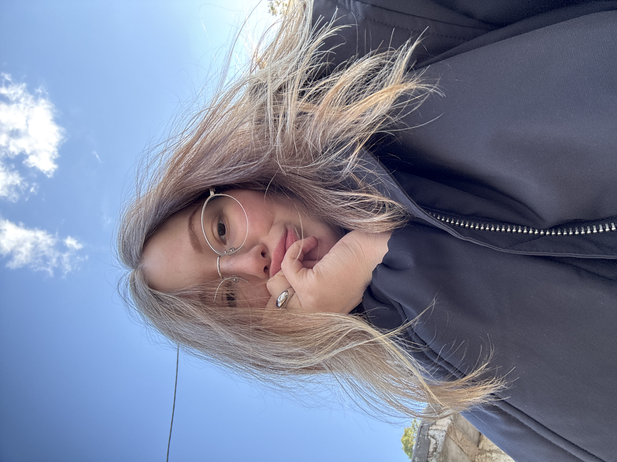 A low-angle, selfie-style image shows a person with light-colored hair, wearing round glasses and a dark jacket, holding their hand to their chin. The background consists of a blue sky with scattered clouds. At the bottom edge of the frame, a partial view of a weathered stone or concrete structure is visible, along with some green foliage.