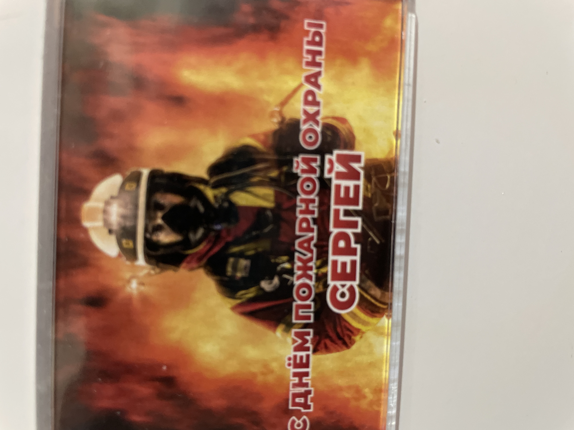 A laminated card or picture shows a firefighter in gear, centered against a fiery background. Vertical Cyrillic text on the right side of the image reads "С Днём Пожарной Охраны Сергей," translating to "Happy Fire Protection Day, Sergey."