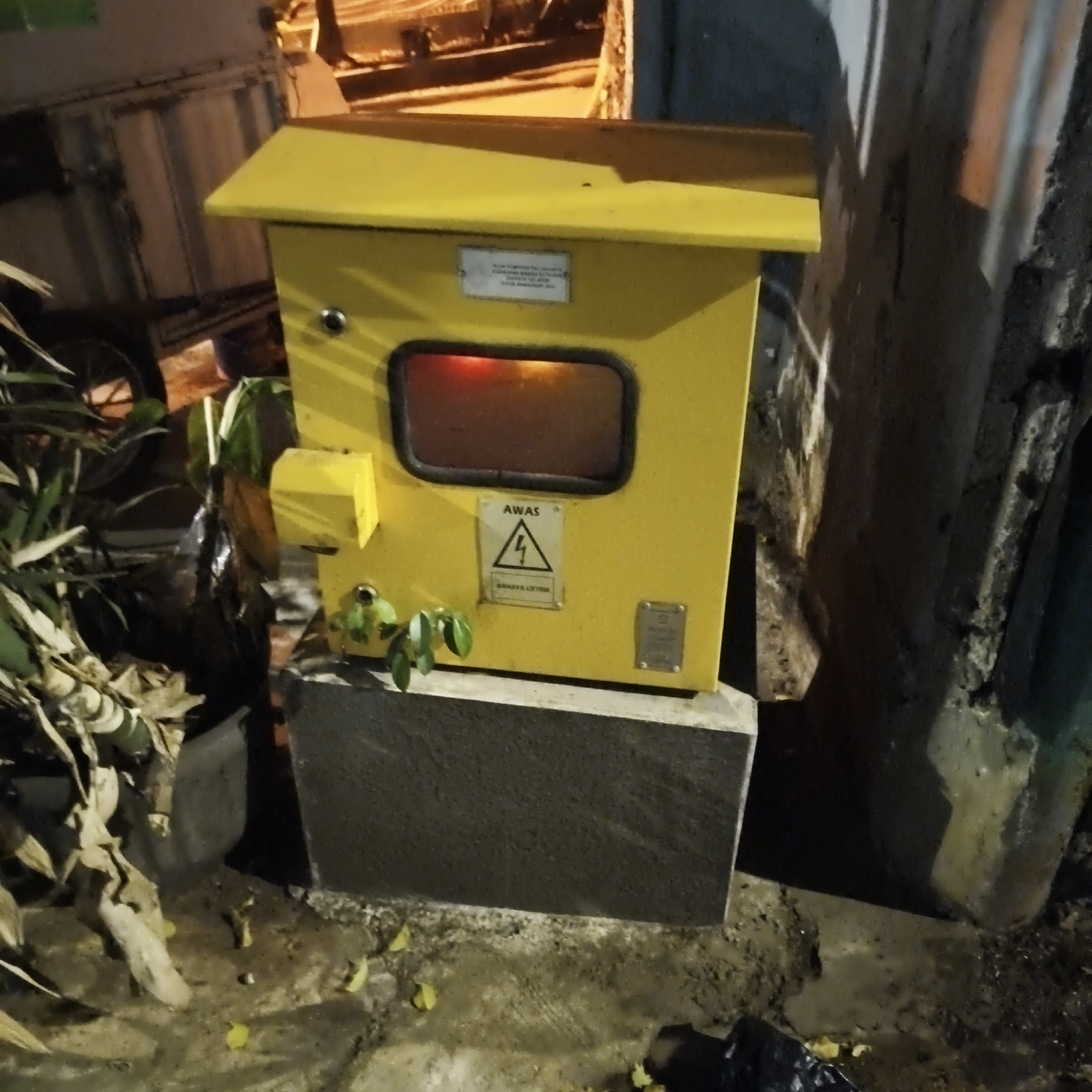 The image depicts a yellow electrical cabinet situated on a concrete base in South Jakarta, Indonesia. The cabinet has a glass-like window with a red glow emanating from within, suggesting it is powered. A caution sign with a lightning bolt symbol and the text "AWAS BAHAYA LISTRIK" is affixed to the front of the cabinet. Surrounding the cabinet is a mixture of dry leaves, green foliage, and some debris. In the background, a large industrial-style container or structure is partially visible, along with a concrete wall and what appears to be a tree trunk. The lighting suggests the photograph was taken at night, with artificial light casting shadows and illuminating parts of the scene. There are no people visible in the image.