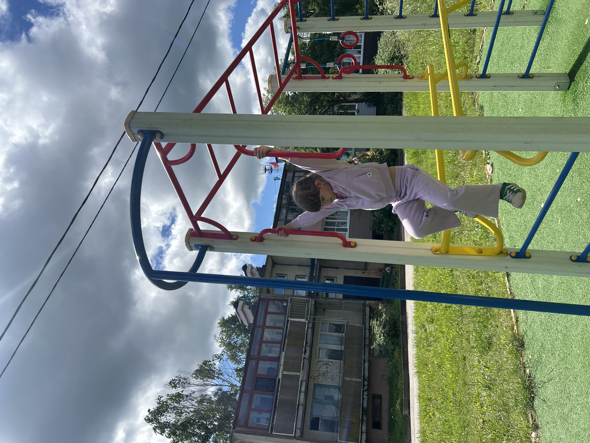 A young girl is climbing on a playground monkey bar structure. She is wearing a light purple tracksuit and sneakers. The playground equipment is made of metal and wood, with blue, red, and yellow accents. In the background, there is a building with several windows and balconies, a green grassy area, and trees. The sky is partly cloudy. The location is identified as Malaya Borshchevka, Russia.