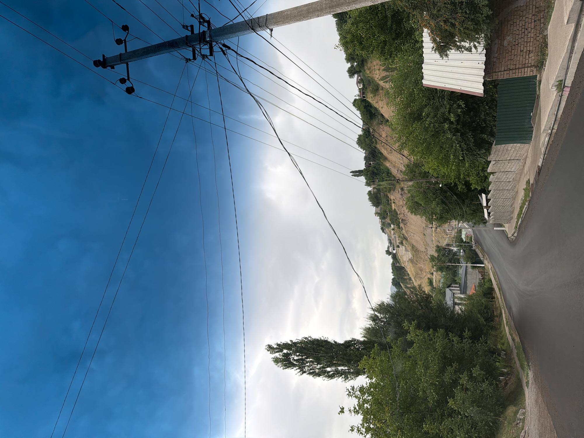 The image depicts a street scene in Khumsan, Uzbekistan. A paved road curves into the distance, flanked by green trees and vegetation on the left and a brick wall with a corrugated metal roof structure on the right. In the background, a steep, dry hillside is dotted with scattered houses and trees. A prominent utility pole with numerous electrical wires is situated on the right side of the road, extending upwards into a bright blue sky with scattered clouds. The overall impression is that of a semi-rural or suburban residential area in a hilly region.