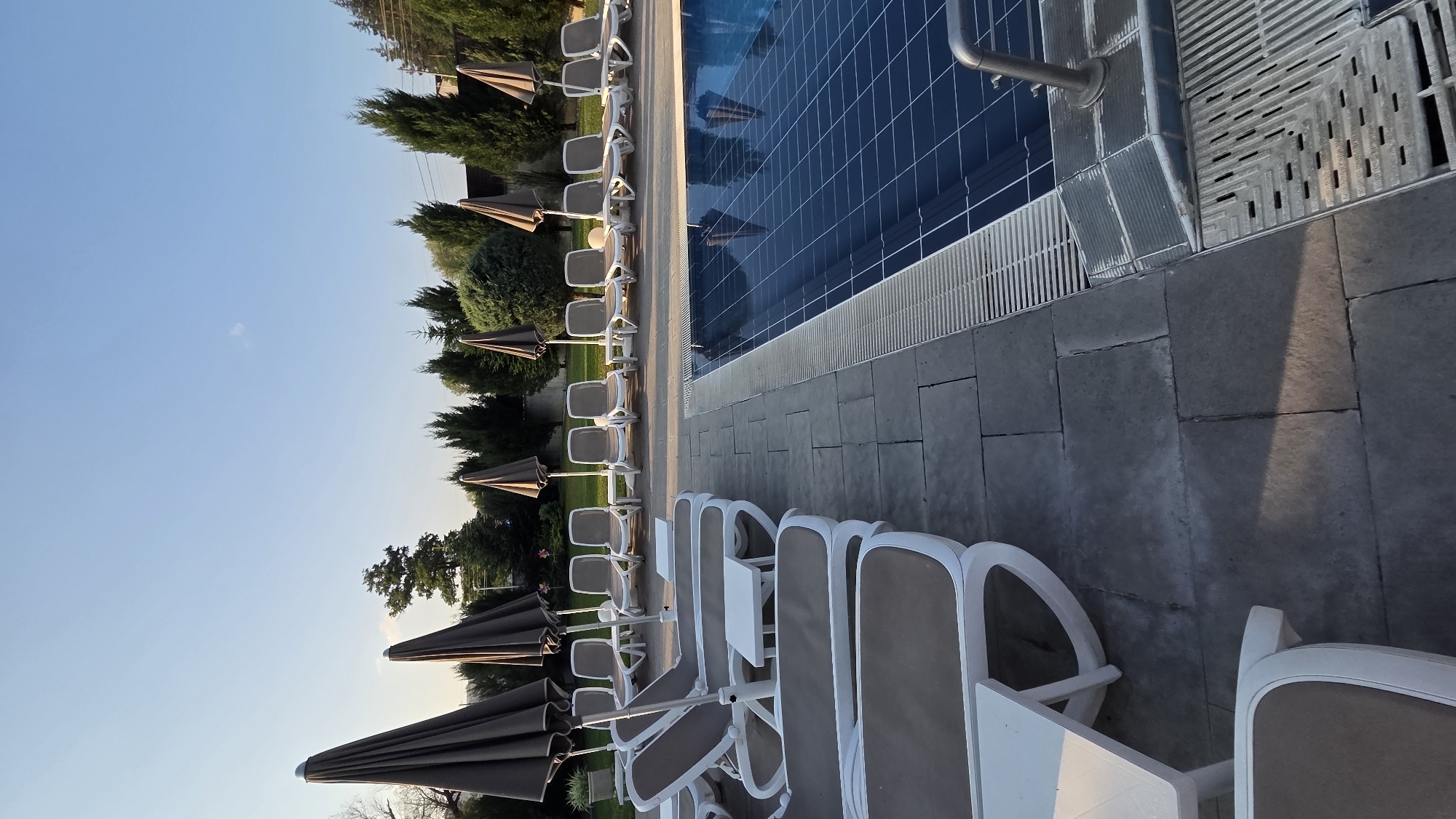 A swimming pool area at dusk features a row of empty, folded-down lounge chairs with brown cushions and white frames, some with small white tables between them. Several closed, dark brown umbrellas stand behind the chairs. The chairs are arranged on a gray tiled patio next to a rectangular swimming pool with a tiled blue bottom. The pool has a metal railing and a white slatted edge. In the background, there is a grassy area with manicured trees and bushes, and a clear blue sky above. The scene appears to be in a resort or hotel setting.