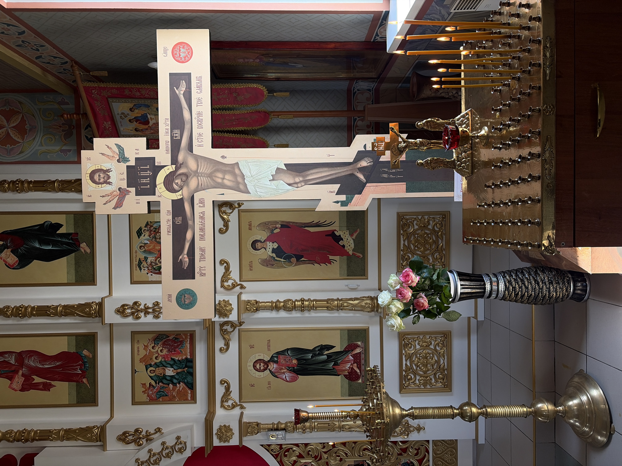 This is an interior shot of a Russian Orthodox Church, likely in Izhevsk, Russia, featuring religious iconography. The central focus is a large paper depiction of the crucifixion of Jesus Christ, adorned with Cyrillic text. Below and around the cross are religious icons, painted in a traditional style. A vase filled with roses sits in front of these icons. To the right is a gold candle stand with numerous lit candles. The walls are painted white with gold ornamentation.