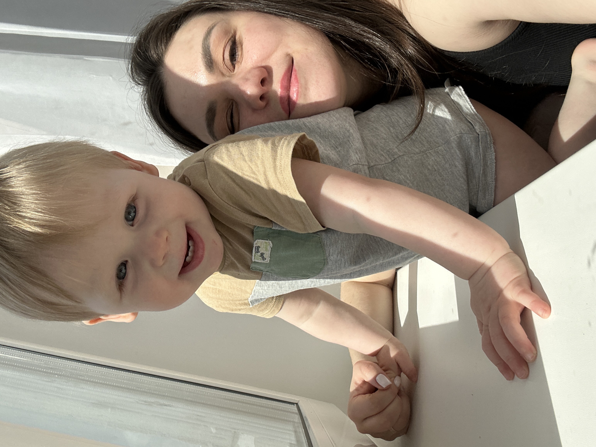 A woman and a young child are posing in an indoor setting, likely in an apartment in the city of Chaikovsky, Russia. The woman has dark brown hair and is wearing a dark tank top. She is lying down, and the child is on top of her. The child has light blonde hair, blue eyes, and is wearing a beige and grey t-shirt with a small green pocket on the front. The child is smiling and looking directly at the camera. They are positioned near a window with a white frame. The scene is illuminated by natural light coming through the window.