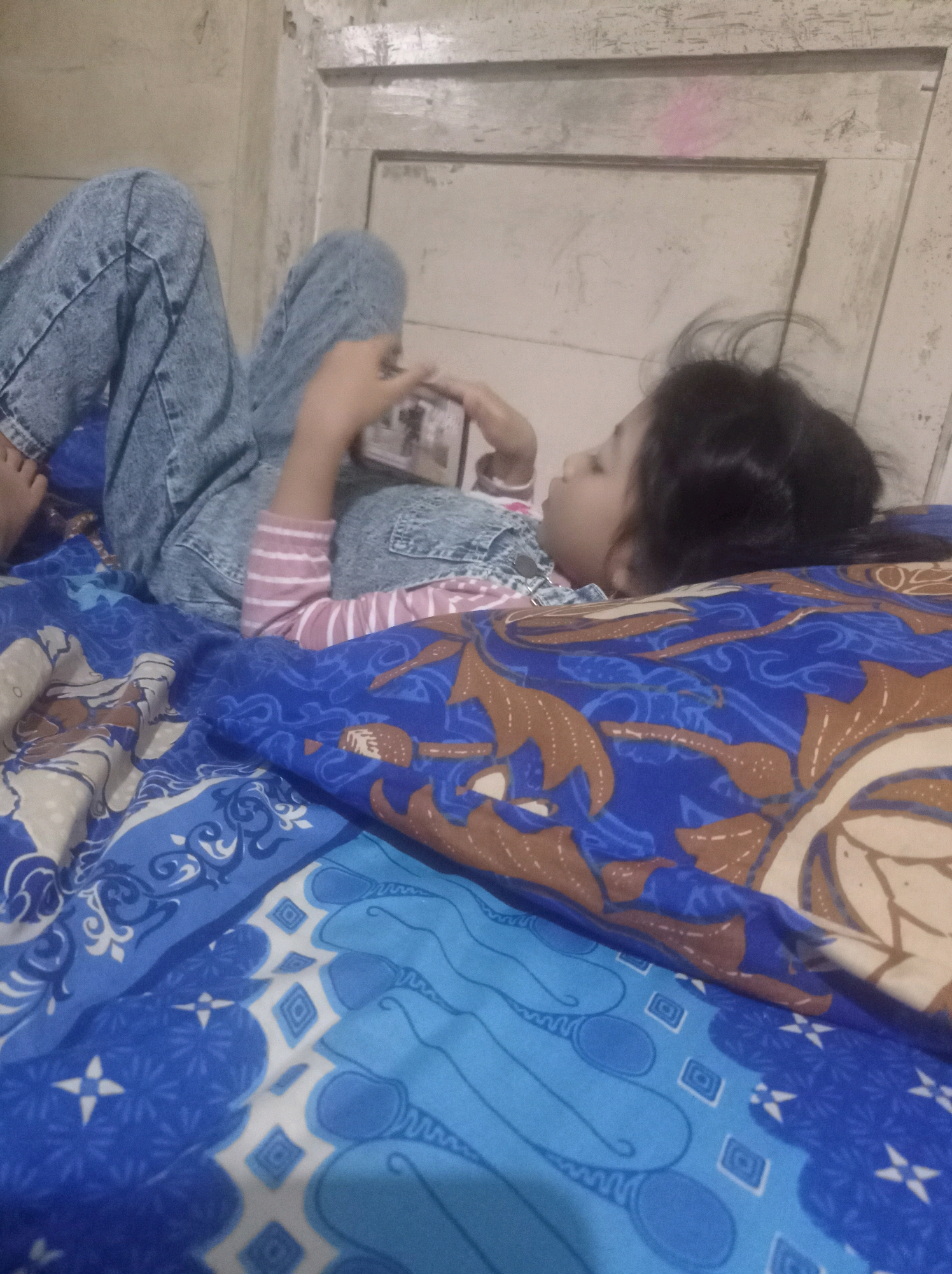 The image shows a young girl lying on a bed, engrossed in her mobile phone. She is wearing light blue denim jeans and a pink and white striped long-sleeved shirt. Her legs are bent, and she is holding the phone horizontally, her hands clearly visible as she interacts with the device. Her hair is dark and appears to be tied back. The bed is covered with a blue patterned sheet, and a pillow with a brown and blue batik design is partially visible. The background suggests an indoor setting with a light-colored wall and a wooden frame, possibly a door or window. A faint pink mark is visible on the wooden frame. The overall lighting is dim, suggesting it might be evening or the room is not well-lit. The girl's posture and focus on the phone indicate she is engaged in a digital activity. The location context is Babalan, Indonesia. There is no visible text in the image.