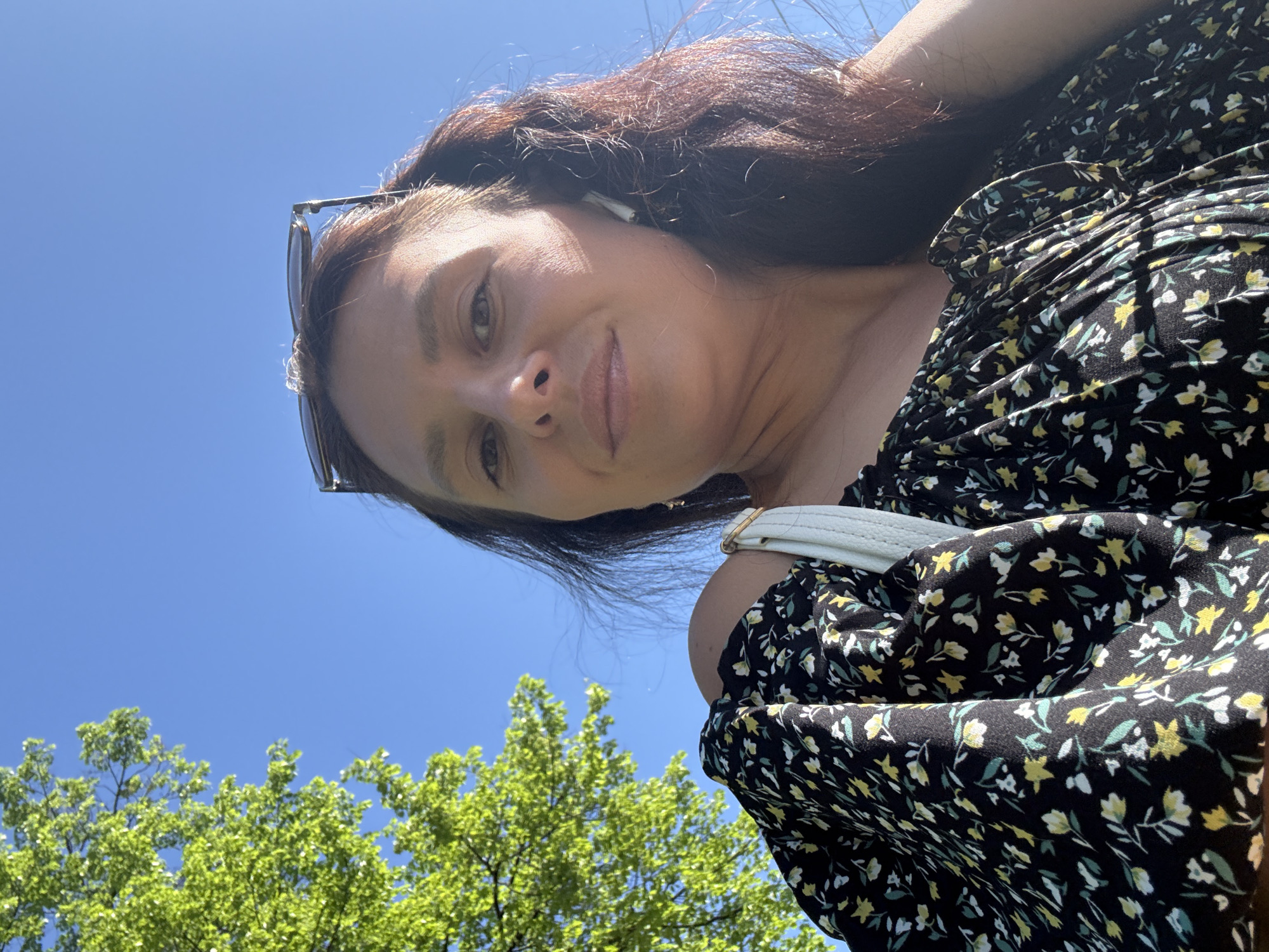 A woman wearing a black floral dress and sunglasses is standing outside. She is looking down at the camera. The background shows a blue sky and green trees. The location could be Chaikovsky, Russia.