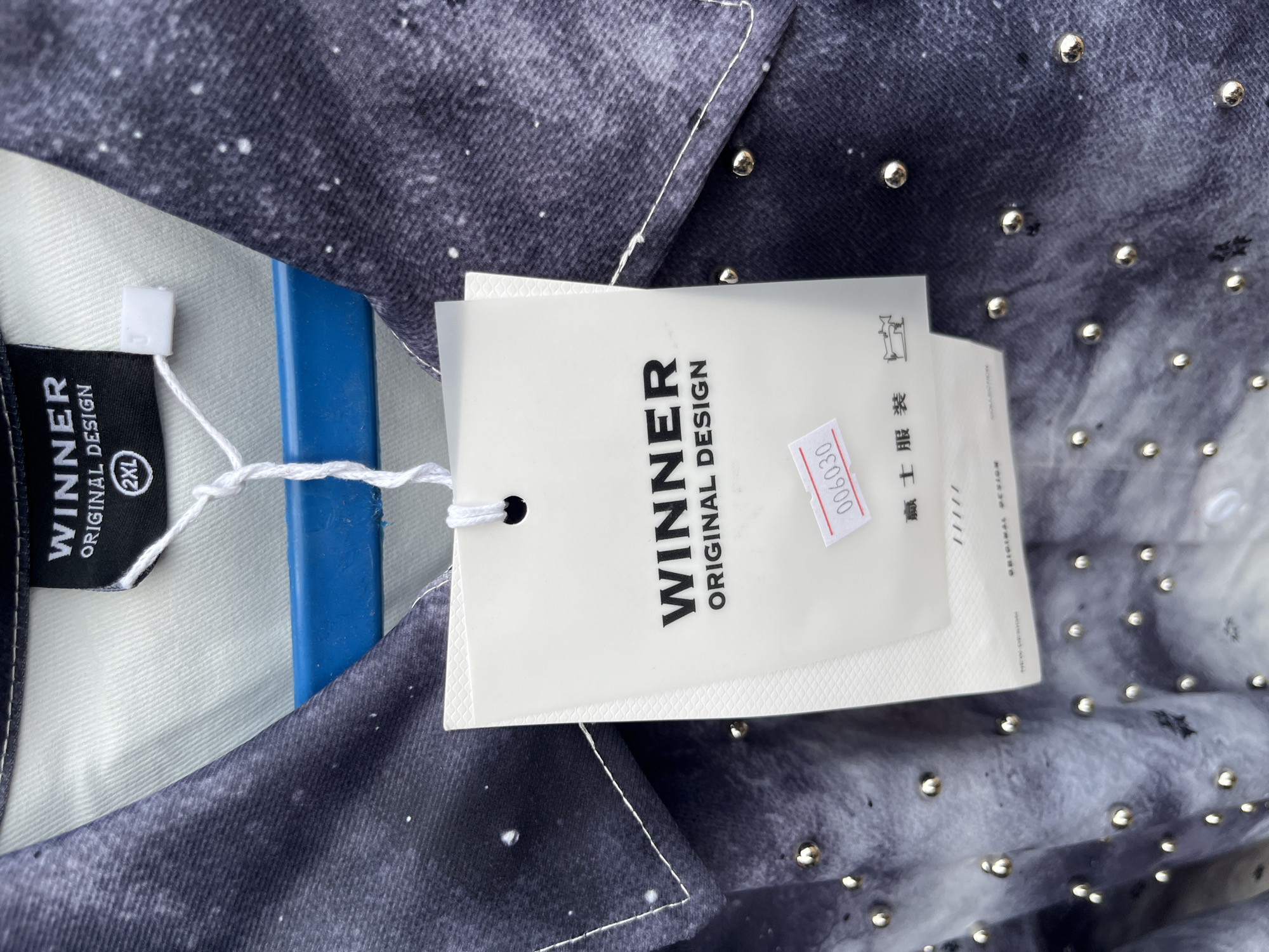 A close-up shot captures a garment with a dark, galaxy-like print, adorned with small, silver studs. A white clothing tag hangs from the garment, displaying the brand "WINNER" in bold black text, followed by "ORIGINAL DESIGN". Below this, a smaller price tag with the number "006030" in red is affixed to the larger tag. The garment appears to be a shirt, possibly denim or a similar fabric, with a collar visible. The background is blurred, suggesting the item is on display, perhaps in a retail setting, or being held up for inspection. The overall impression is one of fashion and branding. There are no people, animals, or other elements to suggest the location is Bukuru, Nigeria.