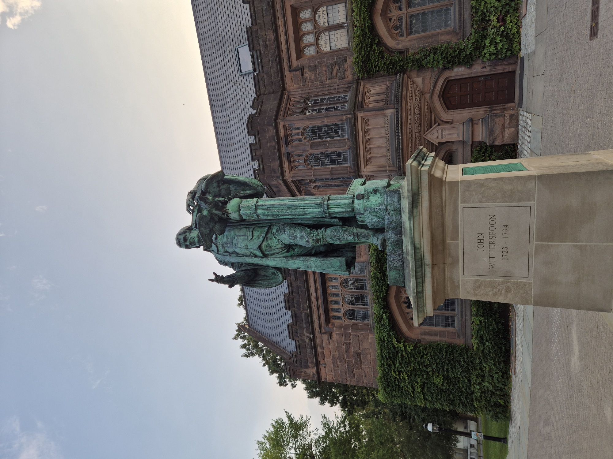A full-shot, low-angle, outdoor photograph captures a weathered bronze statue of John Witherspoon (1723-1794) situated on a stone pedestal in front of a large, Gothic-style stone building with ivy covering portions of its exterior. The statue depicts Witherspoon in period clothing, with his right arm extended upward and his left hand resting on a globe supported by a column. The building behind the statue features arched windows and intricate stone carvings. The sky above is a pale blue with scattered clouds. The ground in front of the statue is paved with interlocking bricks and some concrete. The image appears to have been taken in Princeton, New Jersey, USA, likely on the campus of Princeton University, given the architecture and the presence of the statue of John Witherspoon, a prominent figure in the university's history.