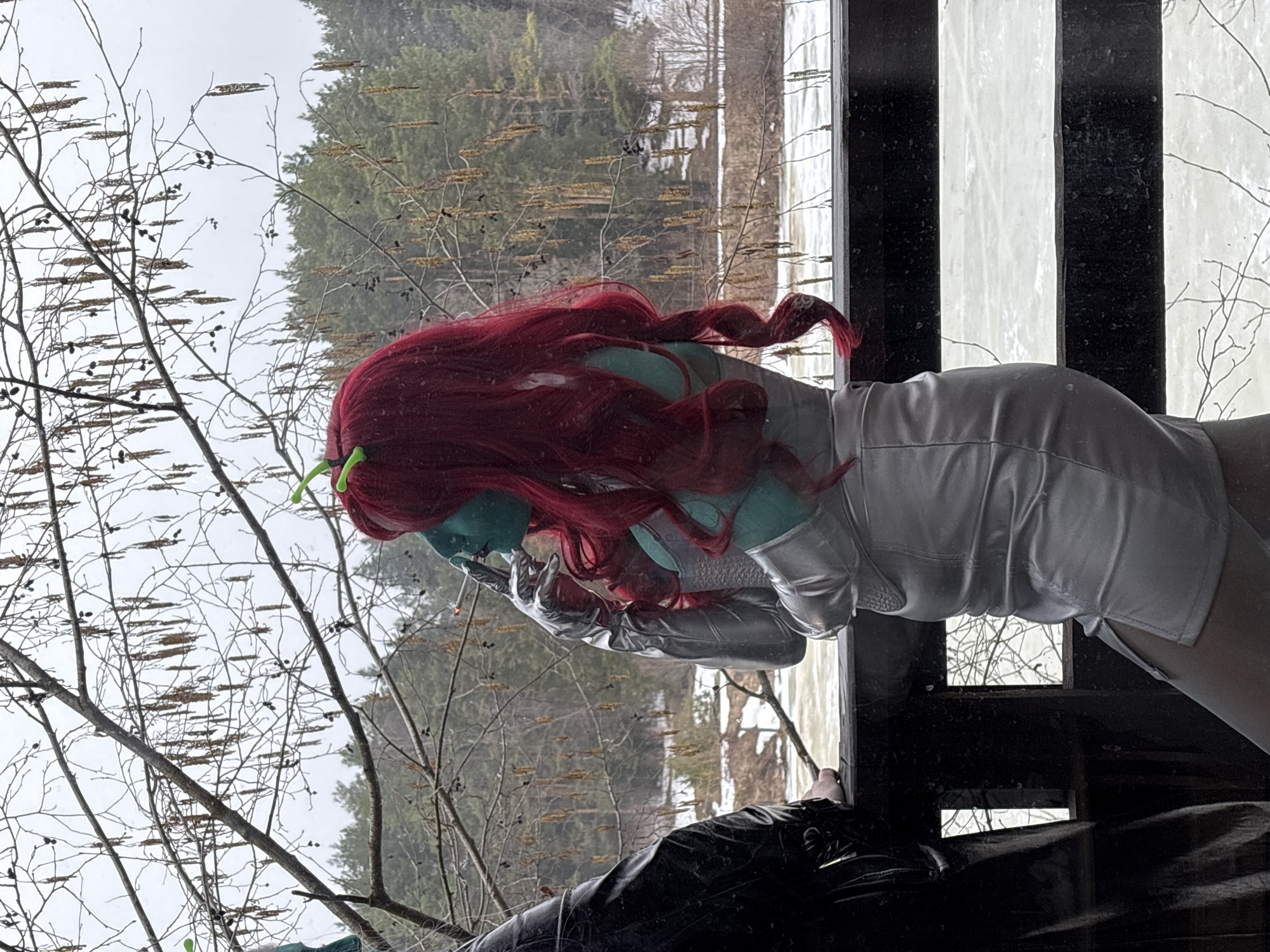 A person with blue skin, red hair, and green antennae is smoking a cigarette. The person is wearing a silver, futuristic-looking outfit with gloves. The person is standing in front of a window or opening that looks out onto a wooded area with bare trees, possibly in a cold or snowy environment. The view through the window suggests a location in a natural or semi-natural setting, possibly near the city of Прикамский, Russia. The scene is depicted in a candid or informal style, focusing on the person and their immediate surroundings.