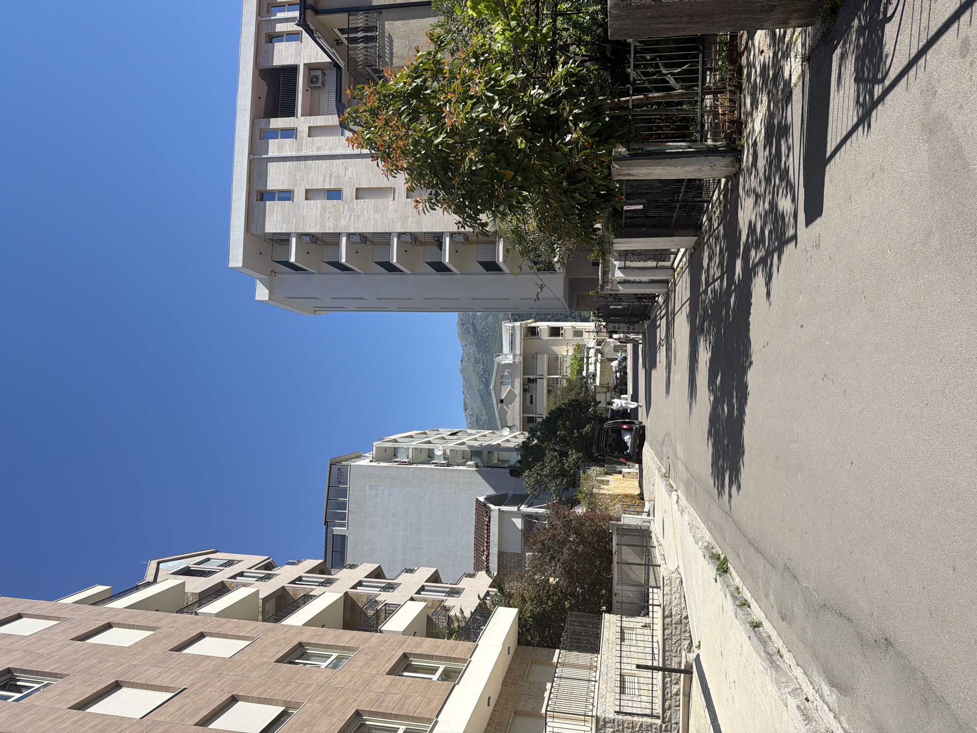 The image displays a sunlit street scene, likely in a town or city, flanked by multi-story buildings. An asphalt road stretches from the foreground into the distance, appearing to follow a gentle incline, with a concrete sidewalk on the right.
On both sides of the street, various buildings rise under a clear, bright blue sky. On the left, facades are predominantly light-colored and modern, featuring multiple balconies, windows, and some visible air conditioning units. A distant building on this side displays a purple sign with the word "SALE" in white letters. On the right side of the street, buildings exhibit a mix of architectural styles, including some with brown-tiled sections. Several mature trees with dense green and reddish-tinged foliage are interspersed among the buildings, casting long, sharp shadows across the sidewalk and road, indicating bright sunshine.
In the mid-ground on the right, a dark-colored SUV or van is parked by the roadside with its rear hatch open. Distant hills or mountains are visible under the clear sky. The scene appears devoid of people, contributing to a calm and quiet atmosphere. The strong, defined shadows suggest it is midday or early afternoon.