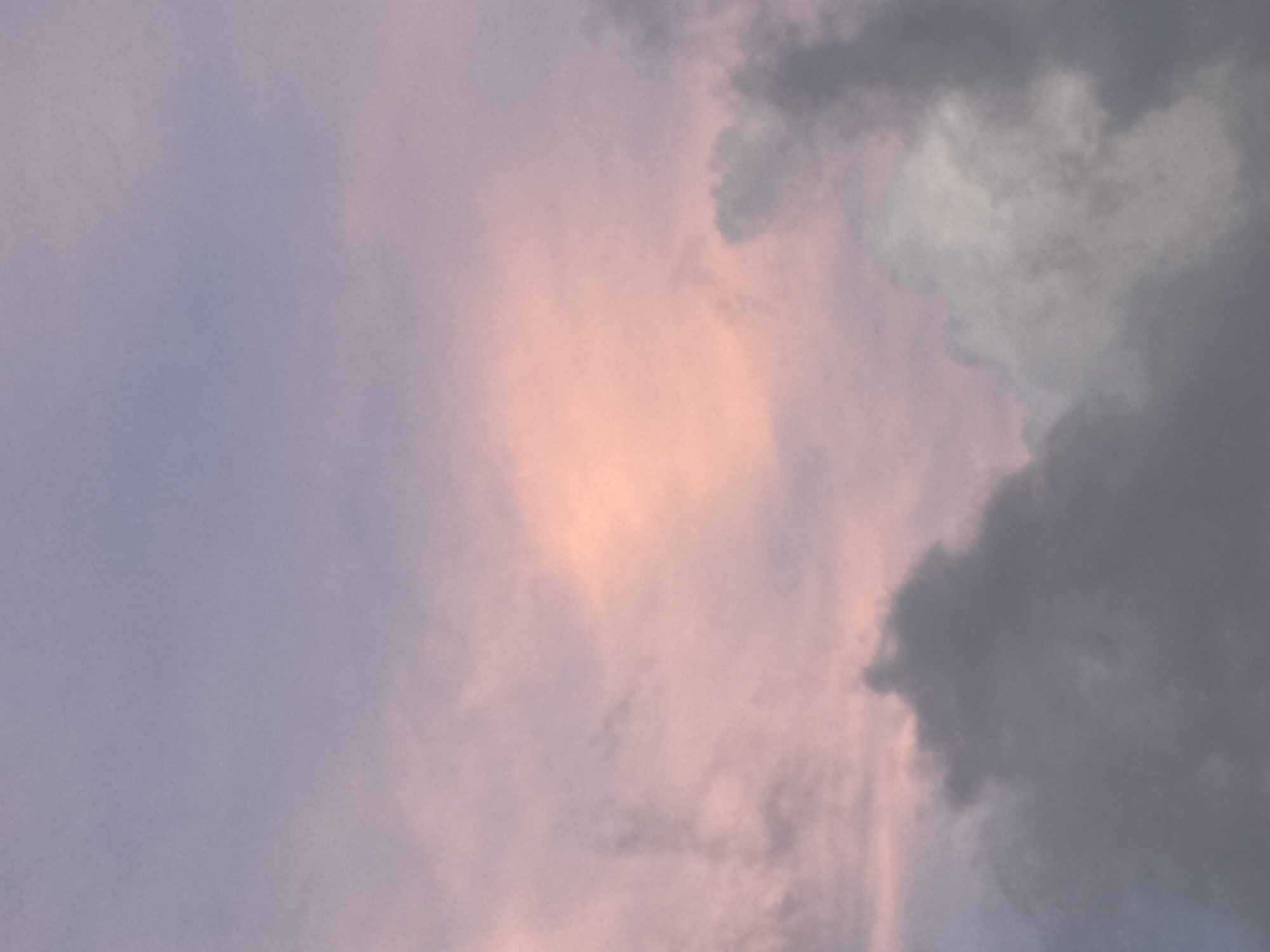 The image depicts a cloudy sky at dusk or dawn in Kuta Selatan, Indonesia. The left side of the frame shows a light purple sky, while the center has a patch of peach or light orange. To the right, there are dark grey storm clouds. There are no people or objects visible in the frame.