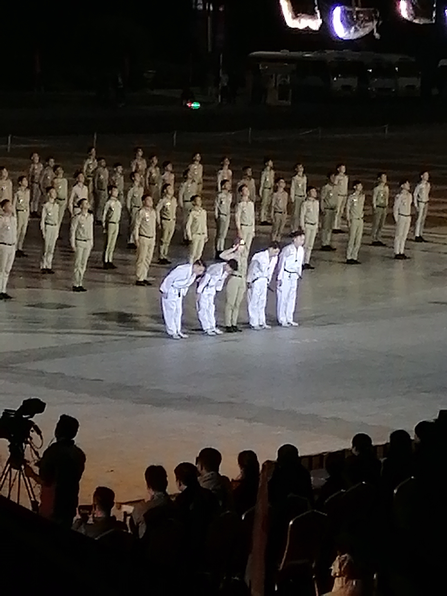 In the city of Da Lat, Vietnam, a group of individuals dressed in matching light-colored uniforms, possibly military or ceremonial attire, are performing a synchronized bow on a large, paved outdoor surface. The individuals are arranged in neat rows, and their posture indicates respect or a formal greeting. In the background, there are blurred figures of other people, some appearing to be spectators or participants in the event, and lighting structures are visible overhead. The scene suggests an organized public event or performance.