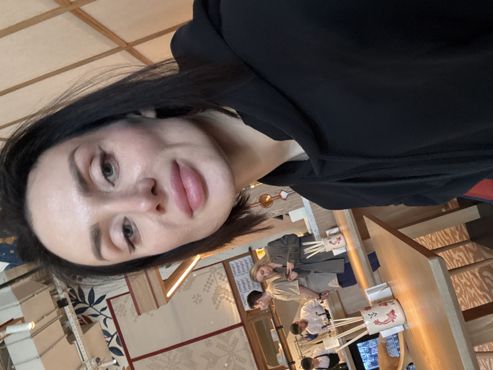 A woman takes a selfie in a brightly lit indoor setting, likely a restaurant or dining area. She is positioned in the foreground, looking directly at the camera with a neutral expression. Her face, characterized by dark eyebrows, winged eyeliner, and full, pinkish lips, occupies the left side of the frame. She has long, dark hair and wears a black hooded top.
The background is slightly out of focus but reveals an active scene. Several individuals are visible behind what appears to be a counter or bar. To the right, a woman with blonde hair in a gray jacket and a man in a light-colored shirt are standing. Further back, two individuals in white chef-like jackets are seen, suggesting a culinary environment.
The setting features light-colored wooden tables or counters, upon which stand cylindrical ceramic holders containing chopsticks. These holders are decorated with red, stylized patterns, possibly depicting a shrimp or fish. Decorative wall panels with intricate geometric and floral designs in blue, white, and brown hues adorn the walls. The ceiling has a grid pattern of light wooden beams. A menu board with unreadable text is also present in the mid-ground. The overall impression is an East Asian-inspired dining or food preparation space during the daytime. No discernable text is visible.