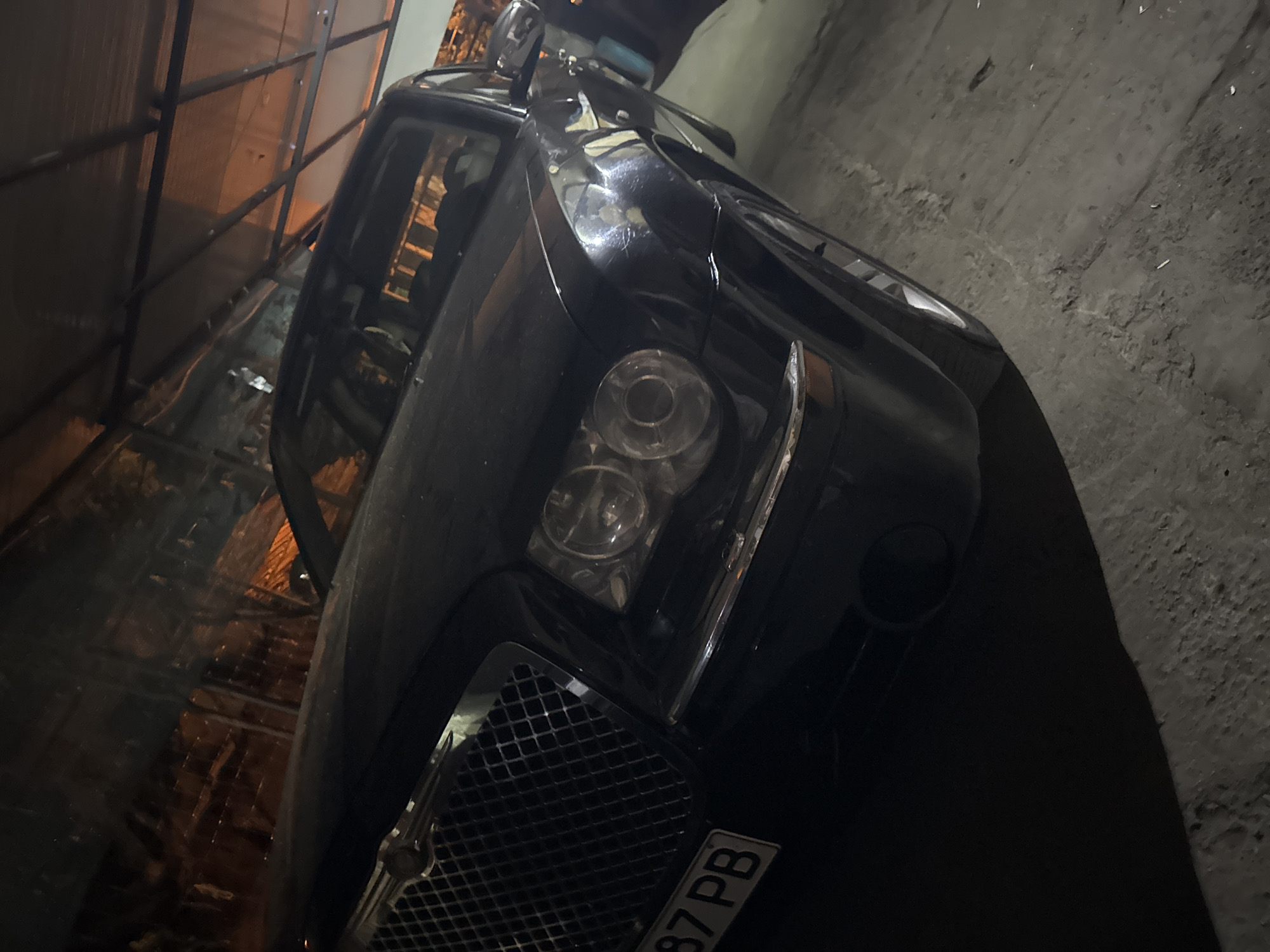 A dark-colored Chrysler 300C sedan is parked in a dimly lit, covered outdoor structure, likely a carport or garage, in Krushare, Bulgaria. The vehicle is angled towards the viewer, showcasing its front left quarter. Its large, chrome-accented grille with a mesh pattern is prominent, topped by the Chrysler emblem. Dual circular headlights with clear lenses are visible on the front left. A partial license plate, displaying "87 PB," is affixed to the lower left of the front bumper. The ground beneath the car is rough and uneven, appearing to be concrete or compacted dirt with scattered debris. Overhead, a metallic frame structure supports a translucent or reflective roof material, from which warm, orange-toned light reflects onto the car's surface and windshield. No individuals are present in the image.
