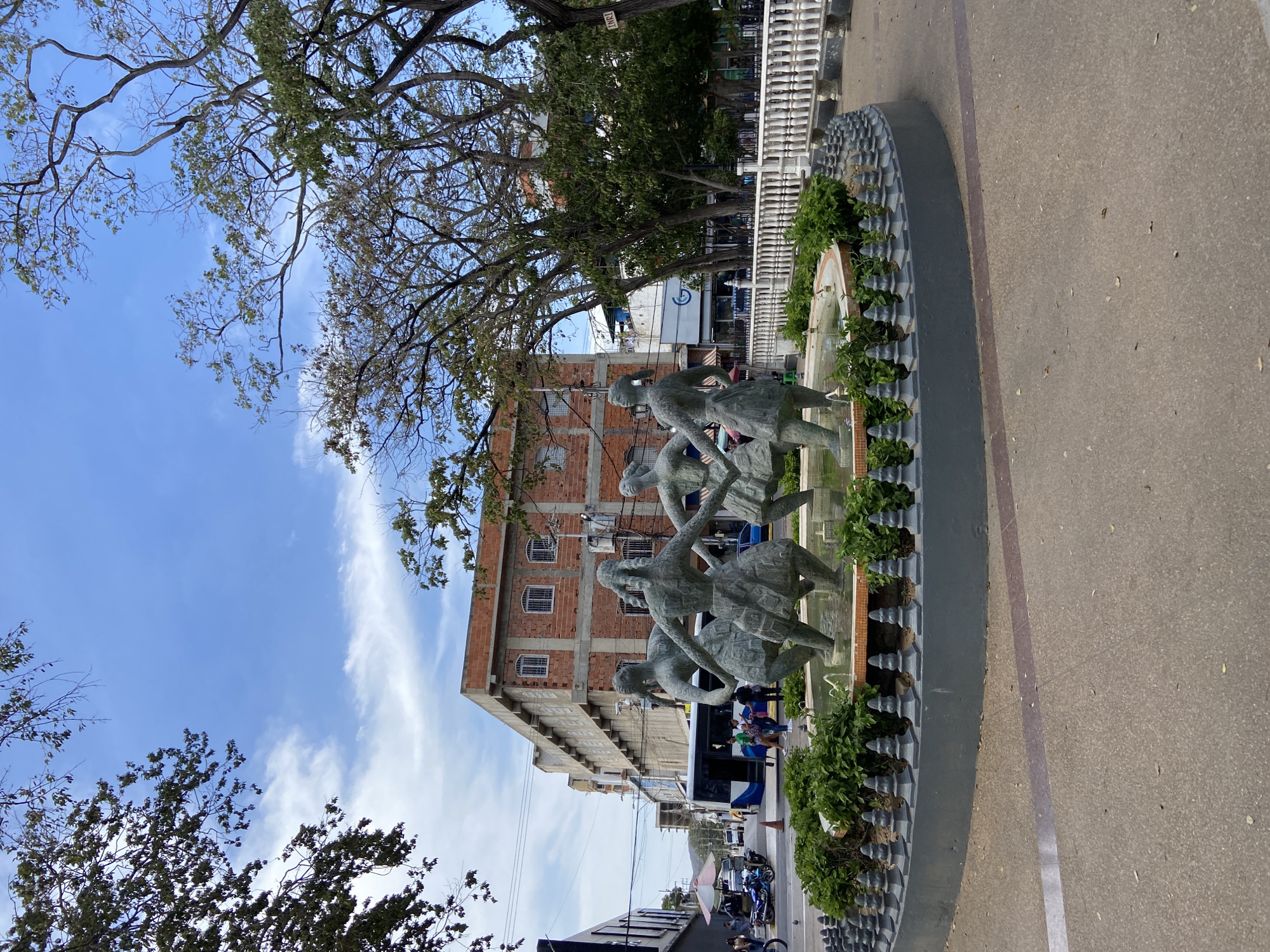 The image depicts a public square in Porlamar, Venezuela, featuring a fountain with sculptures of children dancing. The sculptures are made of a gray material, possibly metal or stone. The fountain is surrounded by a circular planter with green plants. A multi-story brick building is visible in the background. People are present in the background. The sky is blue with some clouds. The image is taken at street level, with a sidewalk in the foreground.