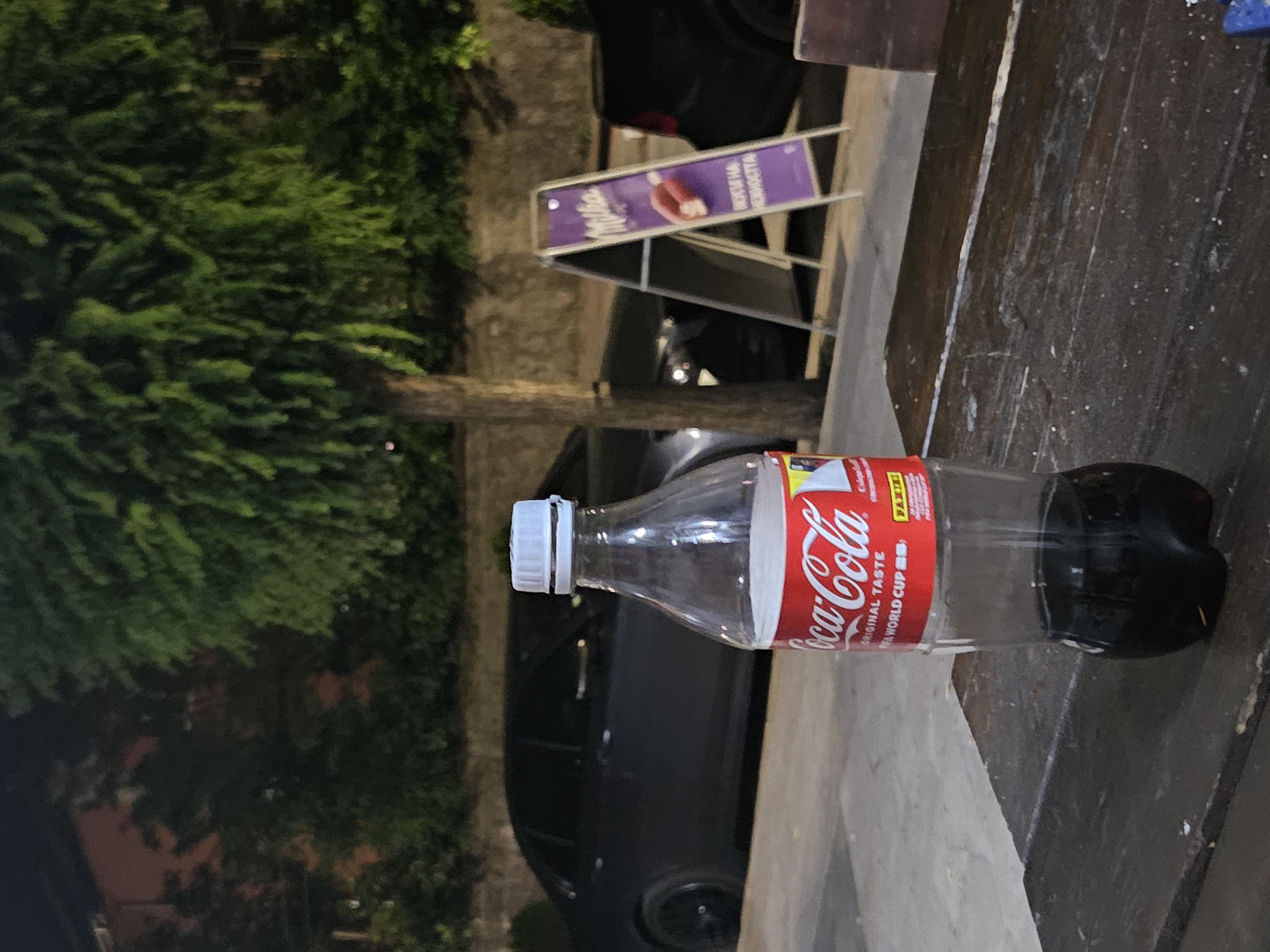 The image shows a plastic bottle of Coca-Cola standing on a wooden table. The bottle is partially filled with a dark liquid, presumably Coca-Cola. In the background, there are cars parked on the street, a tree with green foliage, and a purple A-frame sign advertising Milka chocolate. The scene appears to be outdoors at night, possibly in a city. The overall impression is that of a casual setting, perhaps a cafe or a roadside stop. The location is identified as Biaga, Bulgaria.