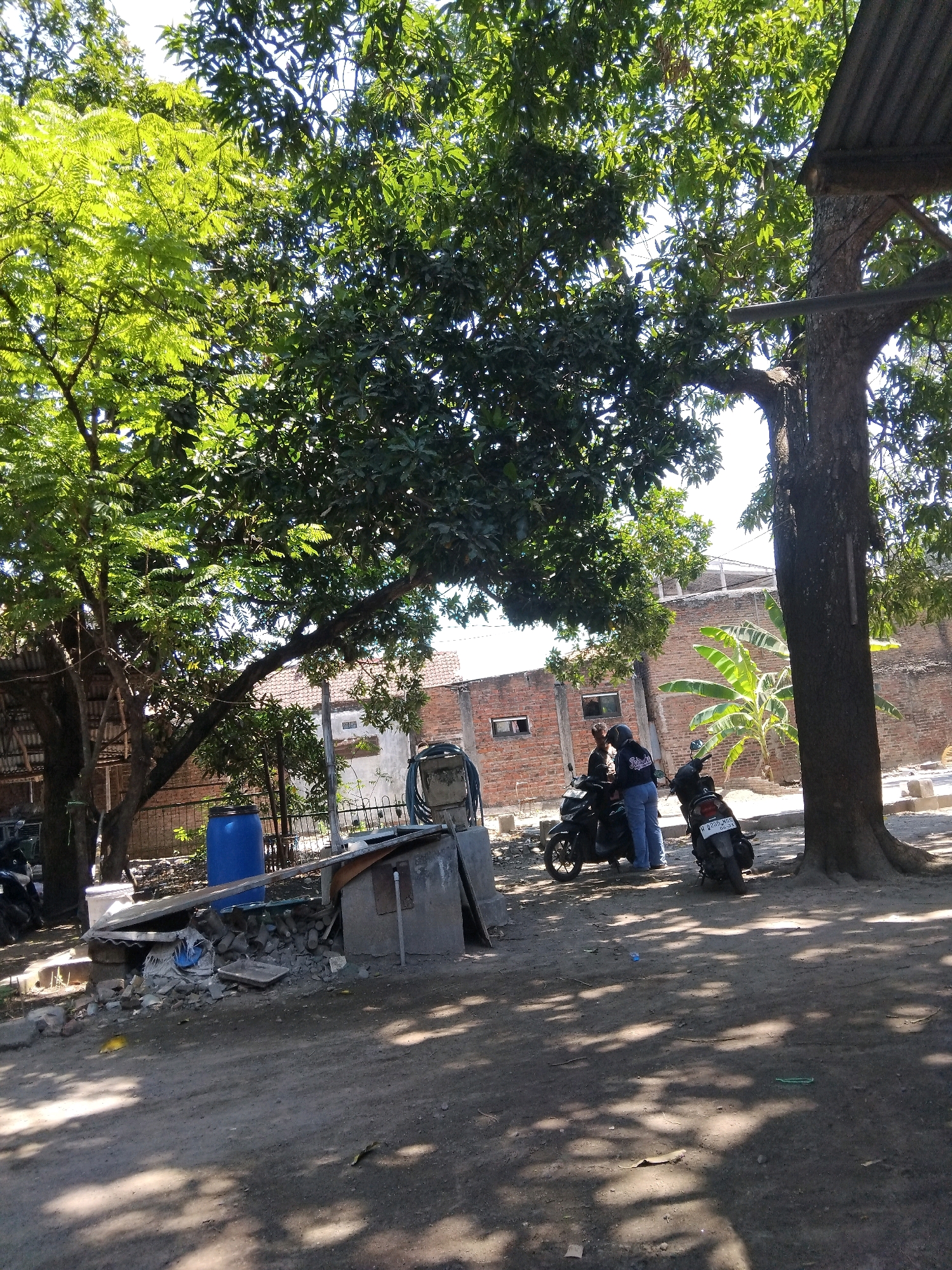 In the city of Kepuh Kiriman, Indonesia, a scene unfolds under the dappled shade of mature trees. Two individuals, one wearing a blue jacket and jeans, and another whose attire is less discernible, are positioned near a black motorcycle. A second, similar motorcycle is parked nearby, its license plate displaying the characters "W 2295 JHC". The ground is unpaved, with areas of packed dirt and scattered debris. To the left, a large blue plastic barrel stands next to a collection of building materials, including corrugated metal sheets and concrete fragments. A brick building with visible window openings forms the backdrop, and a banana tree grows beside it. The overall environment suggests a semi-urban or rural setting with evidence of construction or storage of materials.